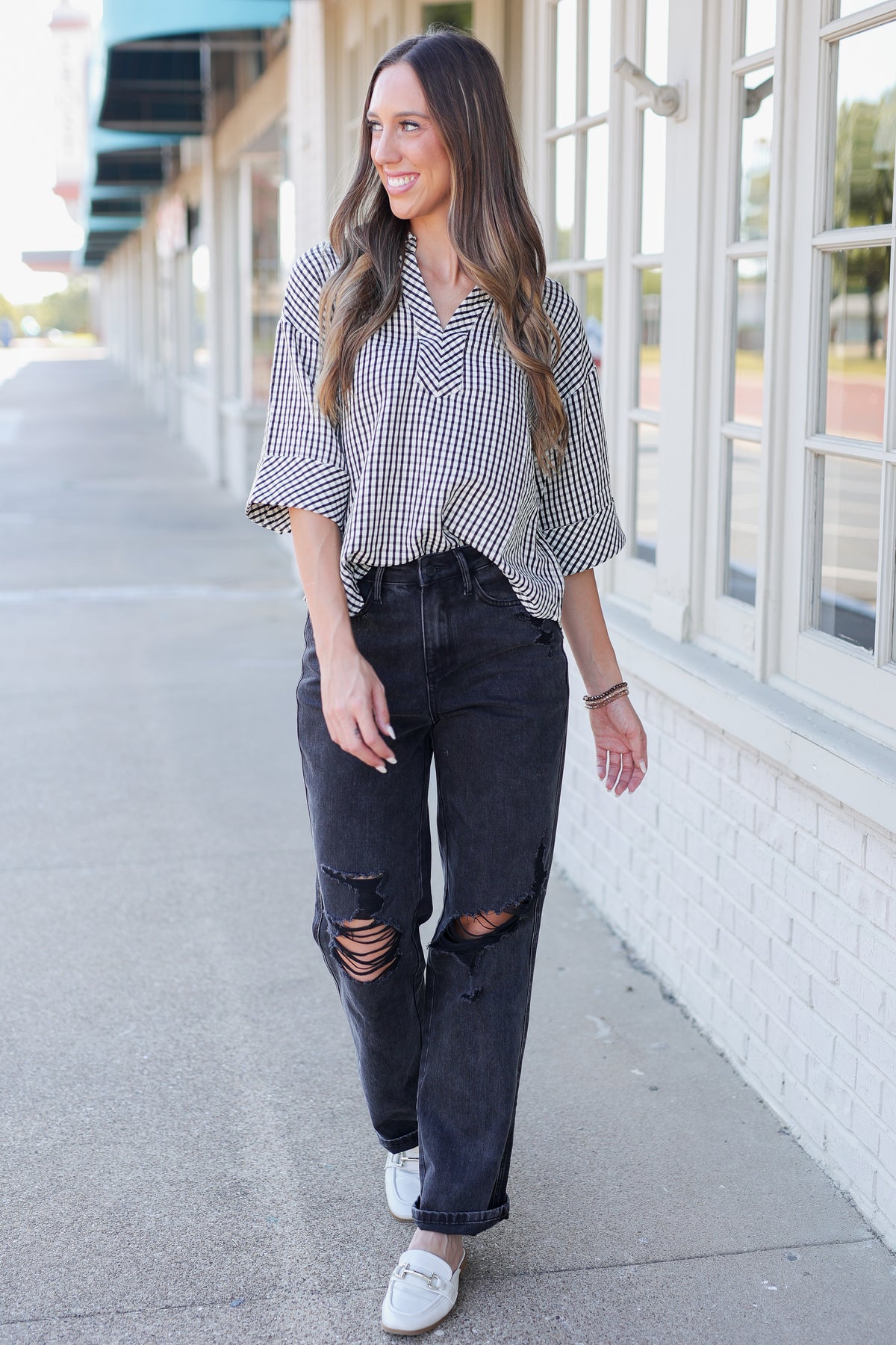 A woman walking outdoors on a sidewalk next to a building with large white-framed windows. She is wearing a black and white gingham button-down shirt, dark gray ripped jeans, and white loafers.
