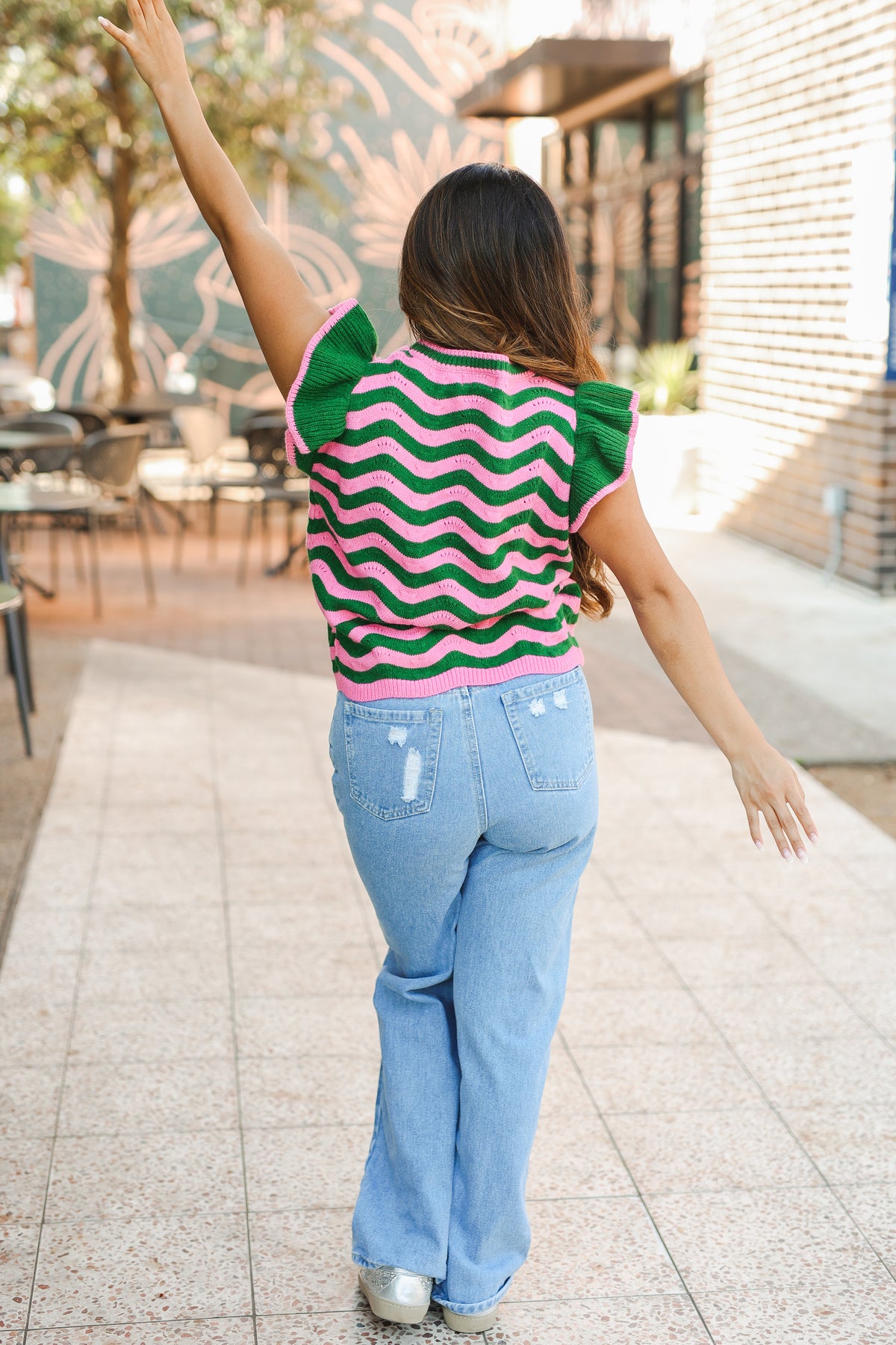 Woman wearing a colorful striped shirt and blue jeans, standing outdoors with one arm raised.