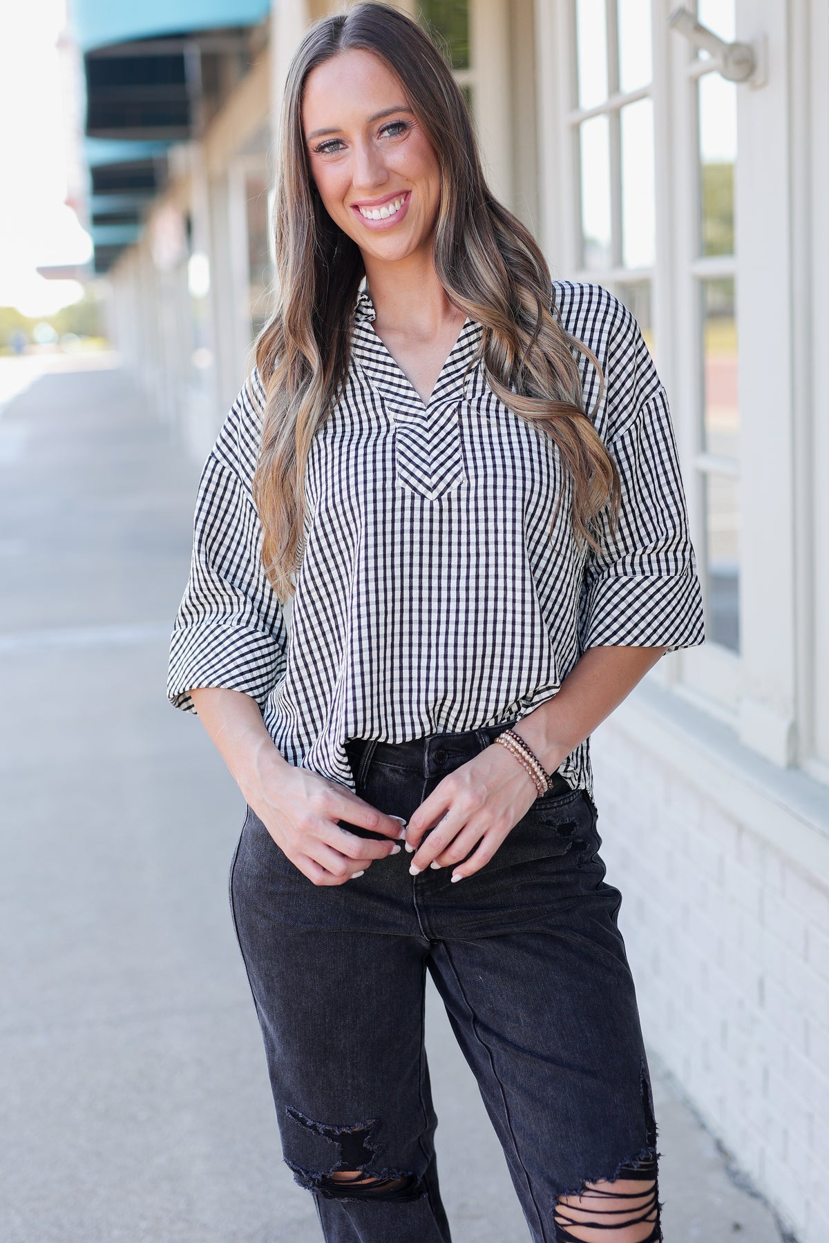 A woman wearing a black and white gingham button-down shirt with rolled sleeves and black distressed jeans, standing outdoors in front of a building with white-framed windows.