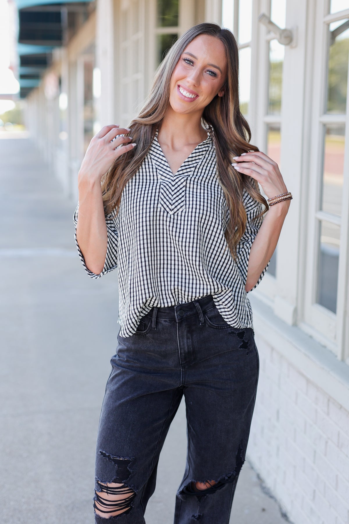 A woman wearing a black and white gingham patterned blouse with three-quarter sleeves and dark distressed jeans, standing outdoors in front of a building with white-framed windows.
