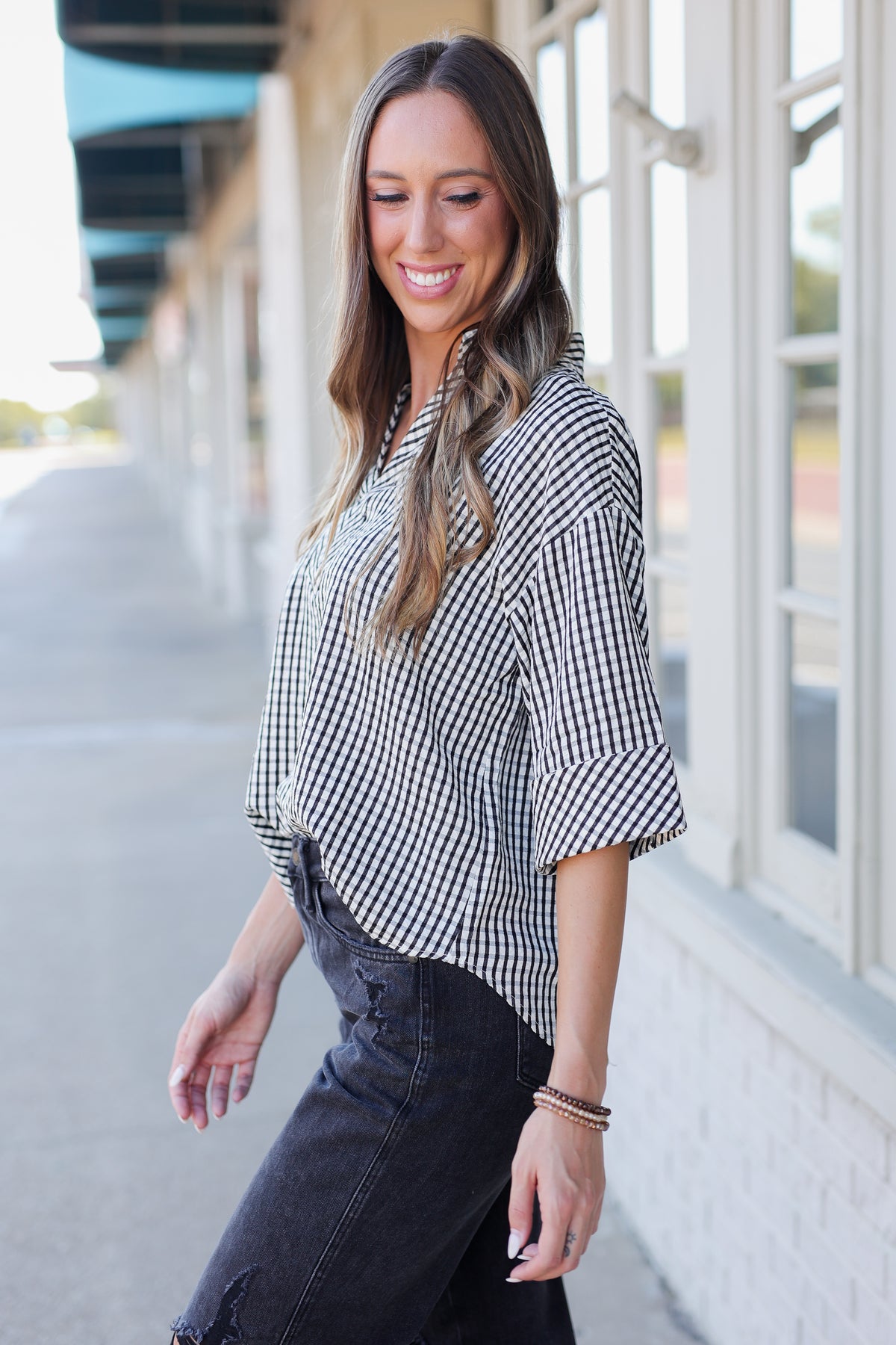 A woman wearing a black and white gingham check short-sleeve blouse and dark denim jeans, standing outdoors in front of a building with large windows.