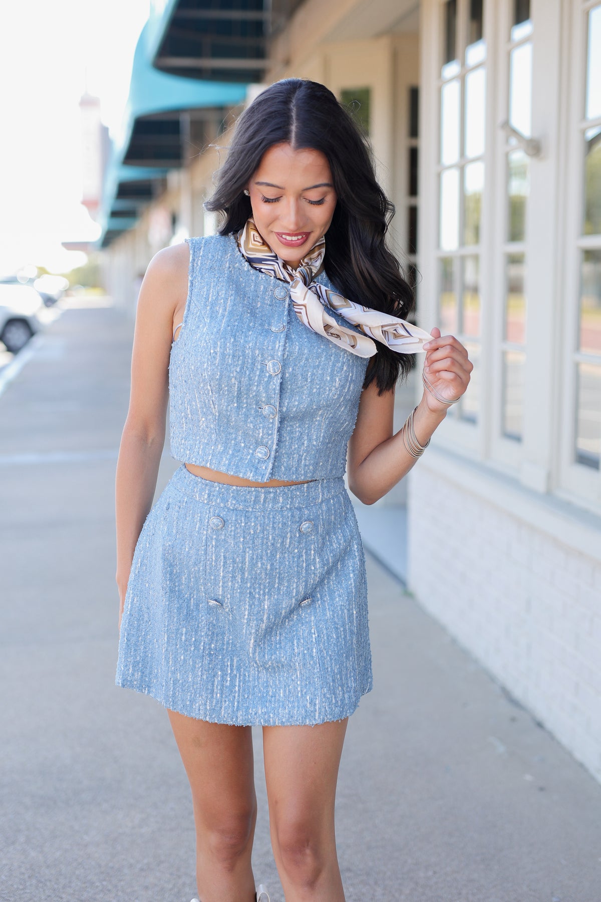 A woman wearing a light blue textured two-piece set consisting of a sleeveless button-front crop top and matching mini skirt, accessorized with a patterned scarf, standing outdoors on a sidewalk.