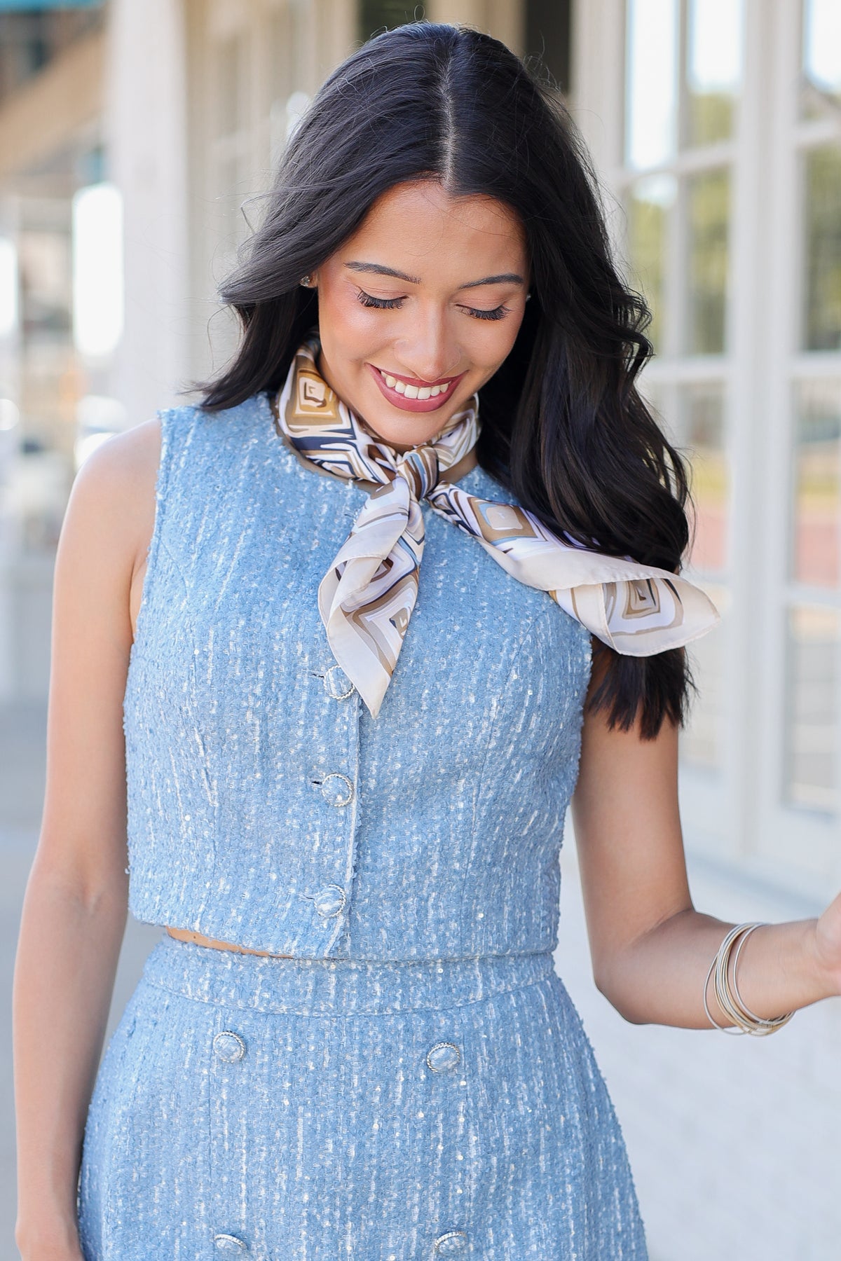 A woman modeling a light blue textured two-piece set consisting of a sleeveless crop top with a patterned neck scarf and a matching mini skirt, paired with white cowboy boots, walking on a sidewalk.