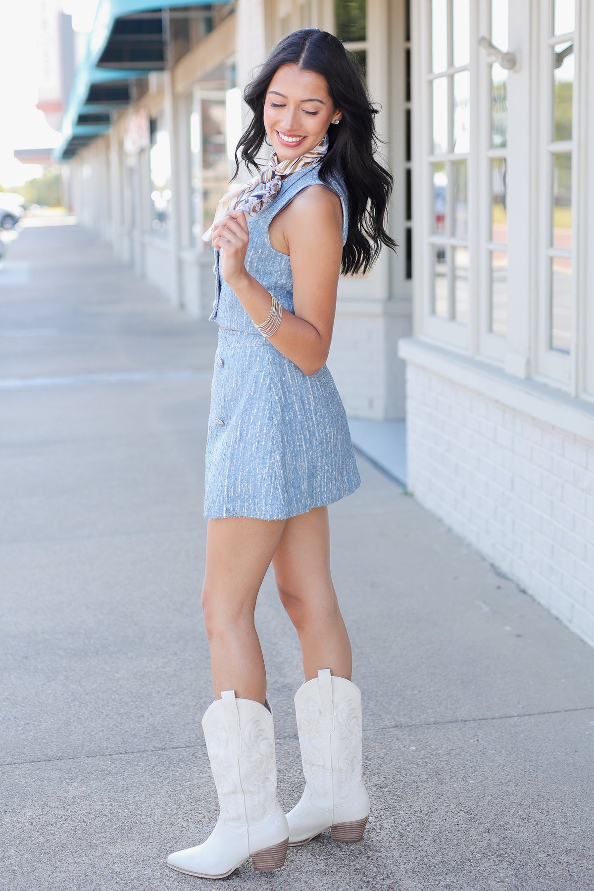 A woman modeling a light blue textured two-piece set consisting of a sleeveless crop top with a patterned neck scarf and a matching mini skirt, paired with white cowboy boots, walking on a sidewalk.