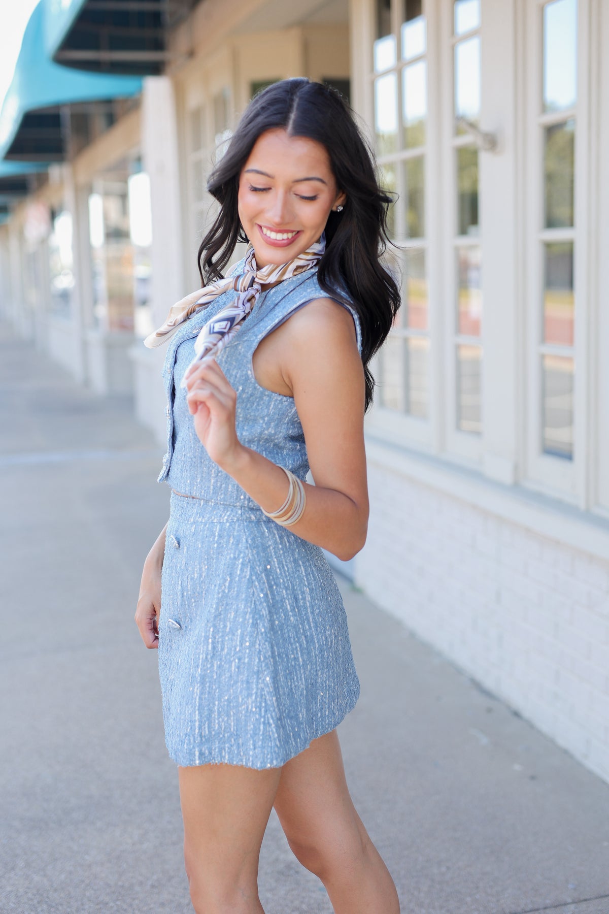 A woman wearing a light blue textured sleeveless vest and matching mini skirt with a patterned scarf, standing outdoors on a sidewalk next to a building with large windows.