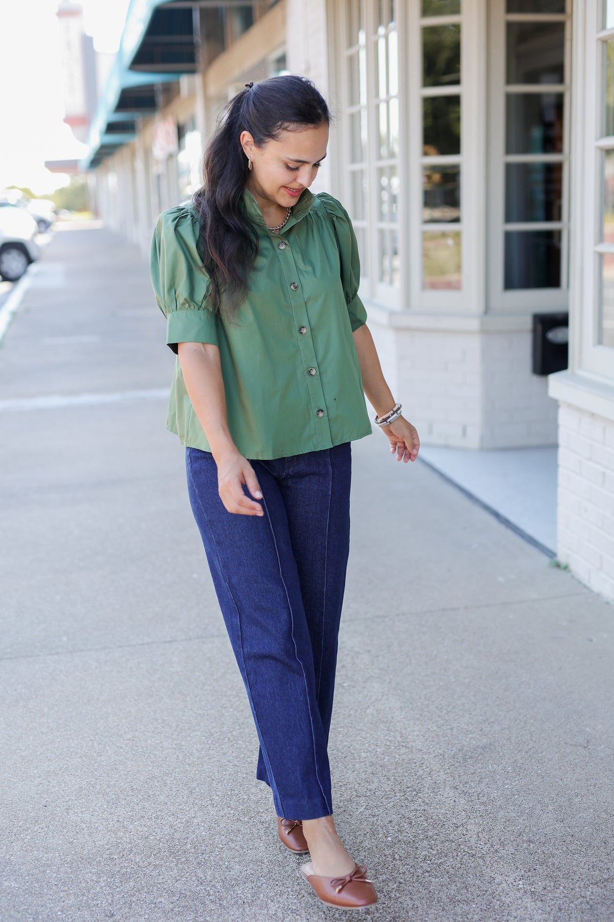 A woman walking outdoors on a sidewalk wearing a sage green short-sleeve blouse with puff sleeves and dark blue wide-leg jeans.