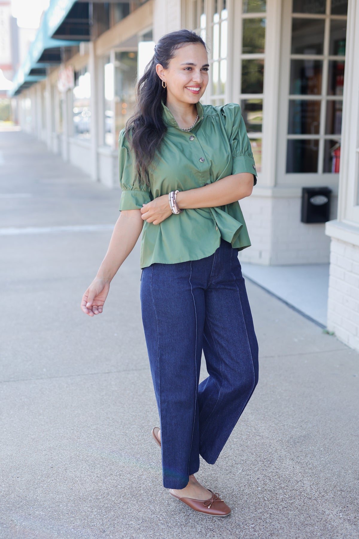 A woman stands on a sidewalk in front of a storefront, wearing a sage green short-sleeve blouse with puffed shoulders and dark blue wide-leg jeans.