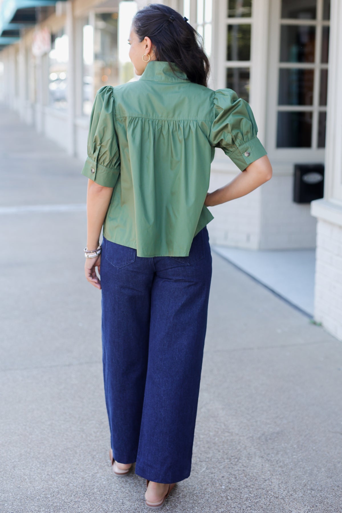 A woman viewed from behind wearing a sage green short-sleeve blouse with puff sleeves and dark blue wide-leg pants, standing on a sidewalk in front of a building.