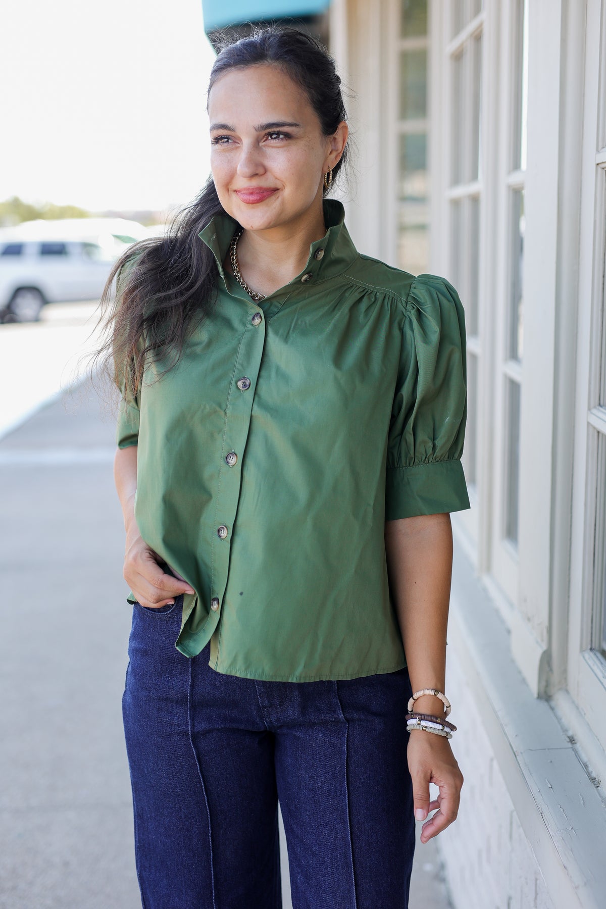 A woman wearing an olive green short-sleeve button-up blouse with puff sleeves and dark blue jeans, standing outdoors next to a building with white-framed windows.