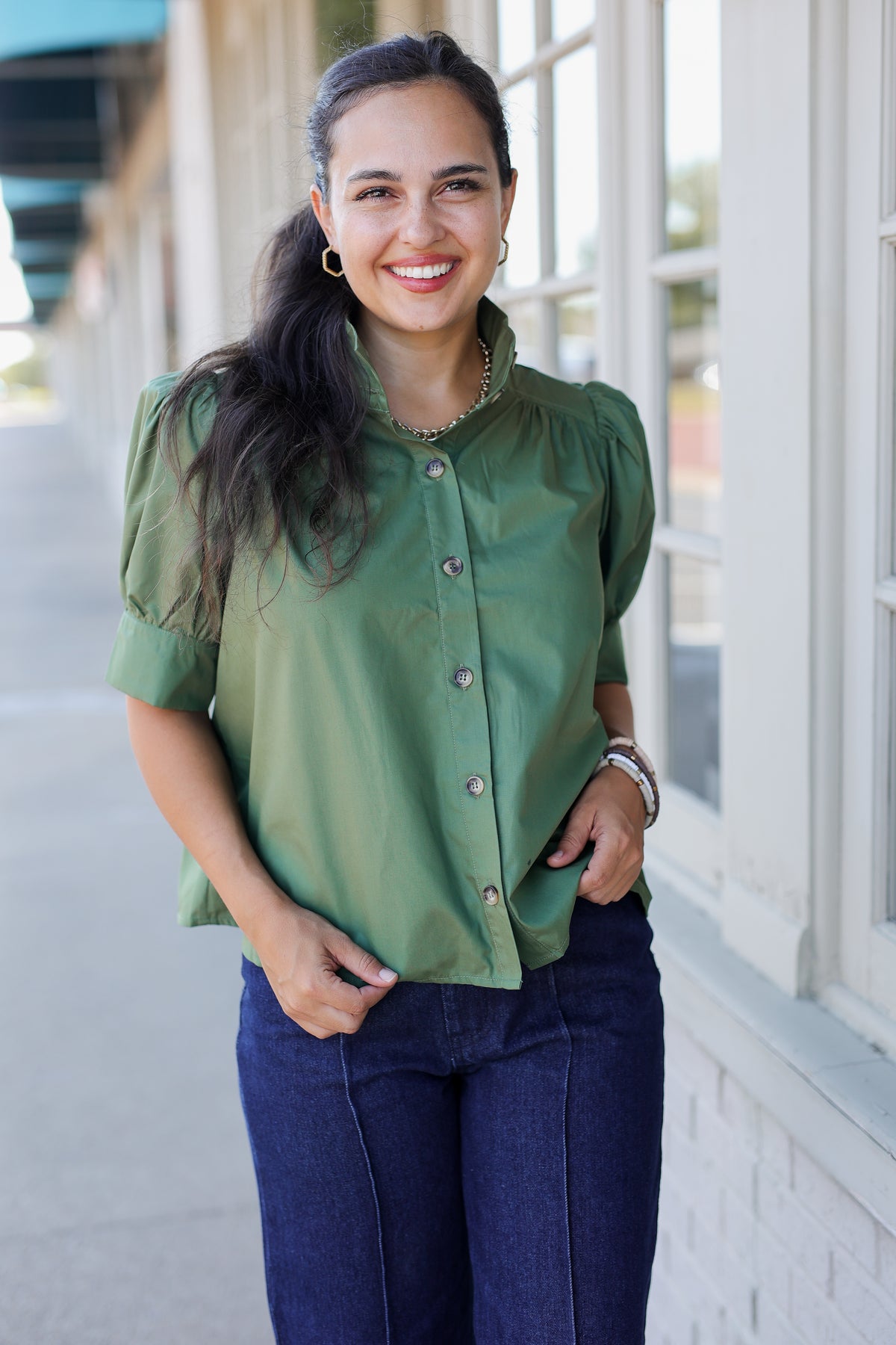 A woman wearing a sage green short-sleeve blouse with puff sleeves and dark blue jeans, standing outdoors in front of a building with white columns and windows.