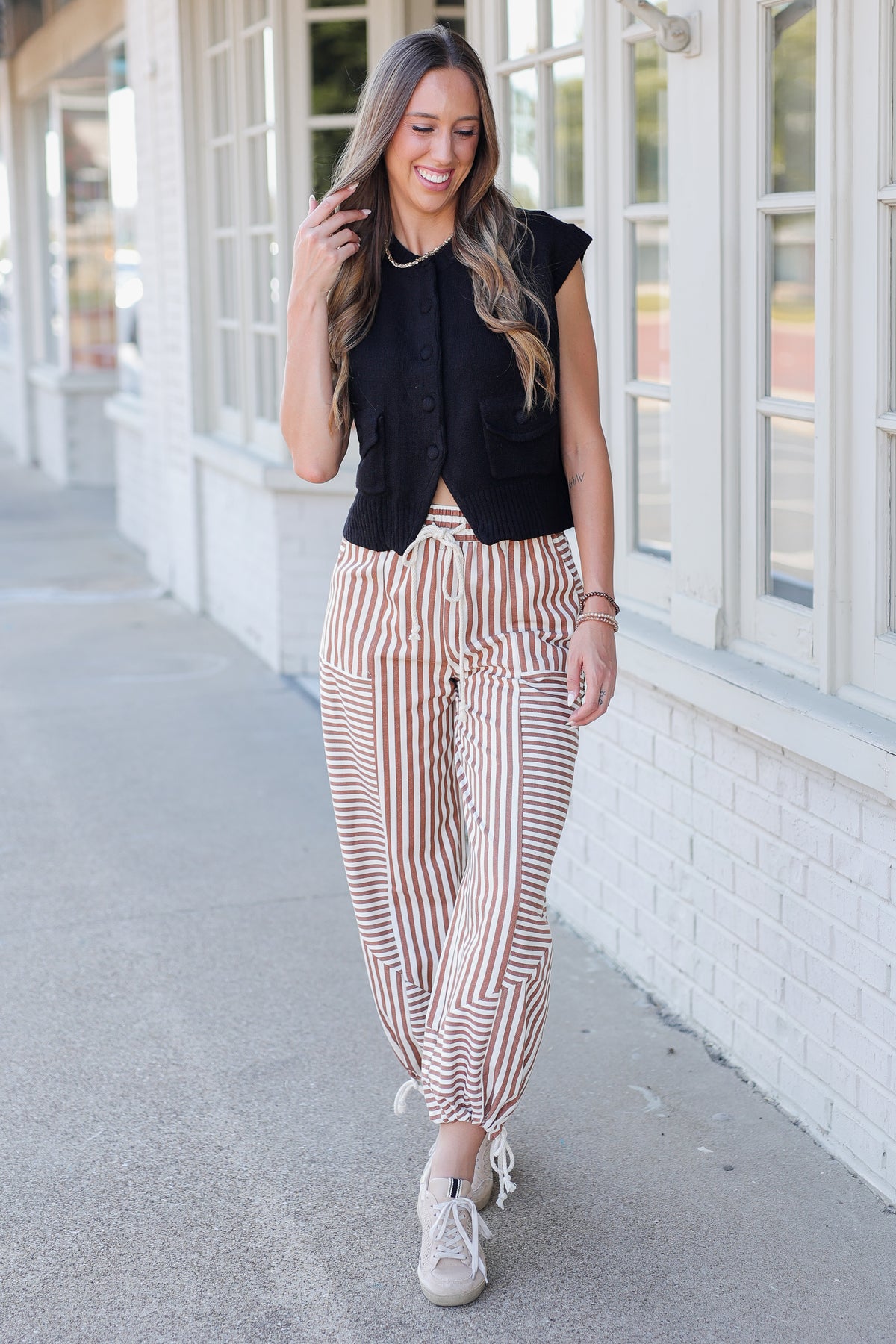 A woman walking outdoors on a sidewalk next to a white brick building, wearing a black sleeveless top, brown and cream striped pants, and white sneakers.