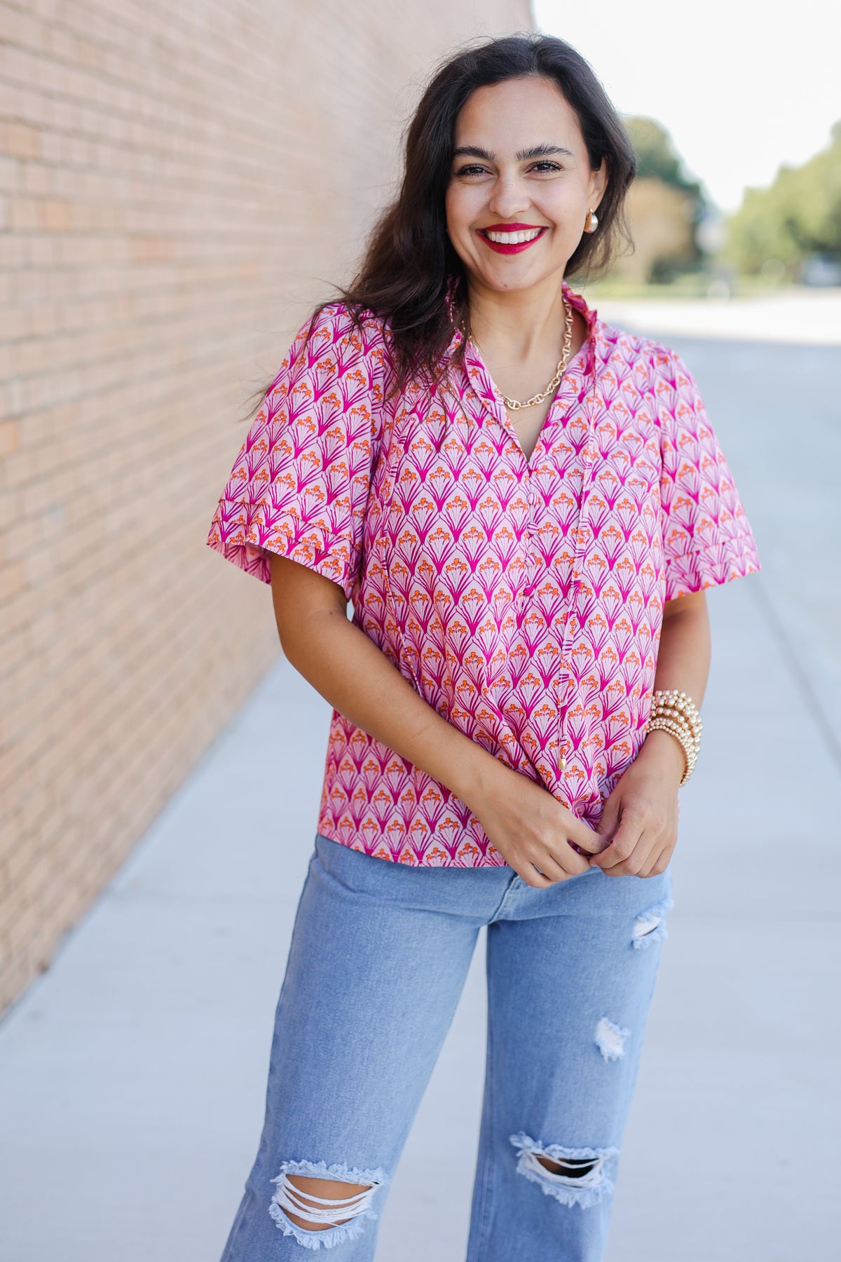 Woman wearing a pink patterned blouse and blue jeans standing outdoors.