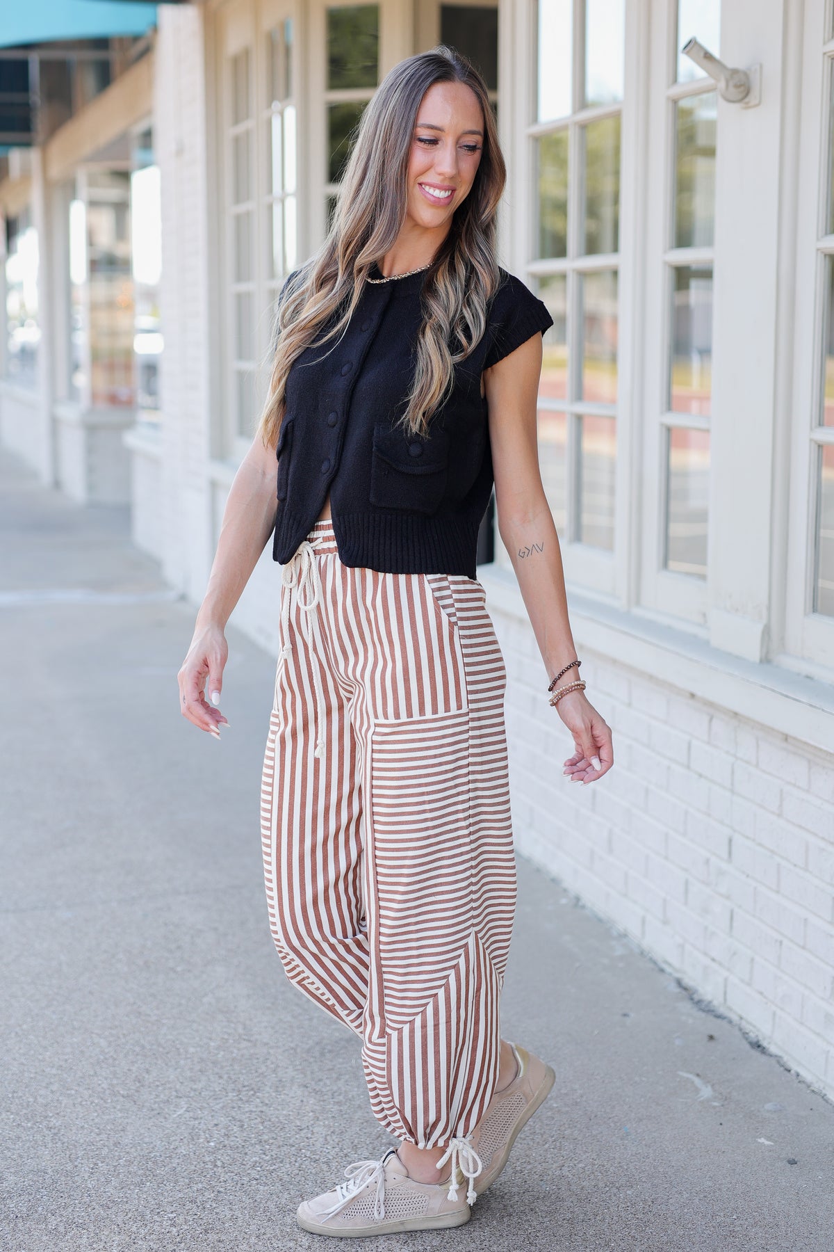 A woman walking outdoors on a sidewalk next to a white brick building, wearing a black sleeveless top, brown and white vertically striped wide-leg pants, and beige sneakers.