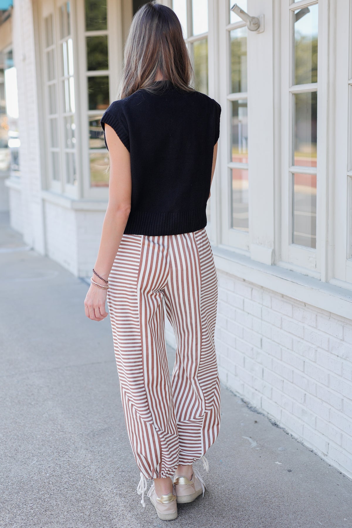 A woman viewed from behind wearing a black sleeveless top and striped wide-leg pants with white fringe at the hems, standing on a sidewalk next to a white brick building.