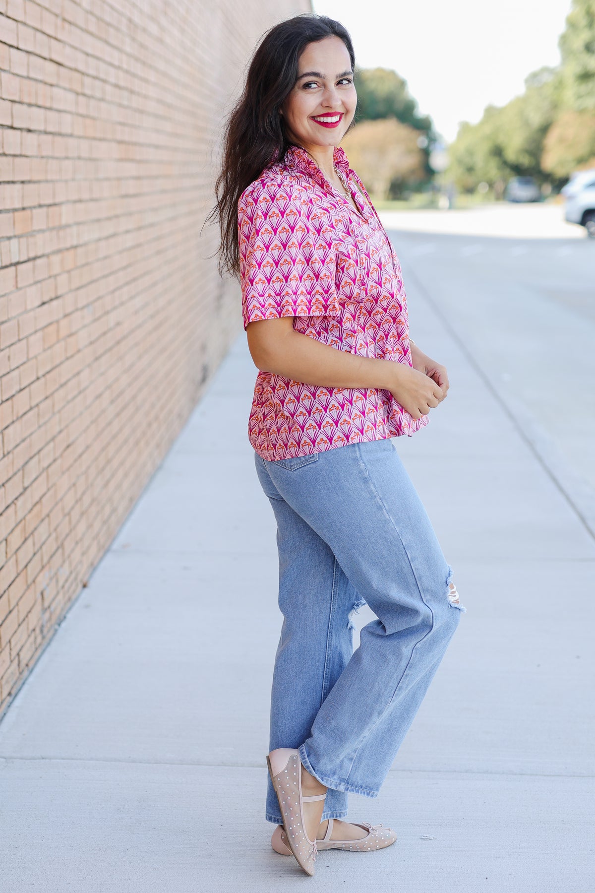 Woman wearing a pink patterned shirt and blue jeans standing against a brick wall.