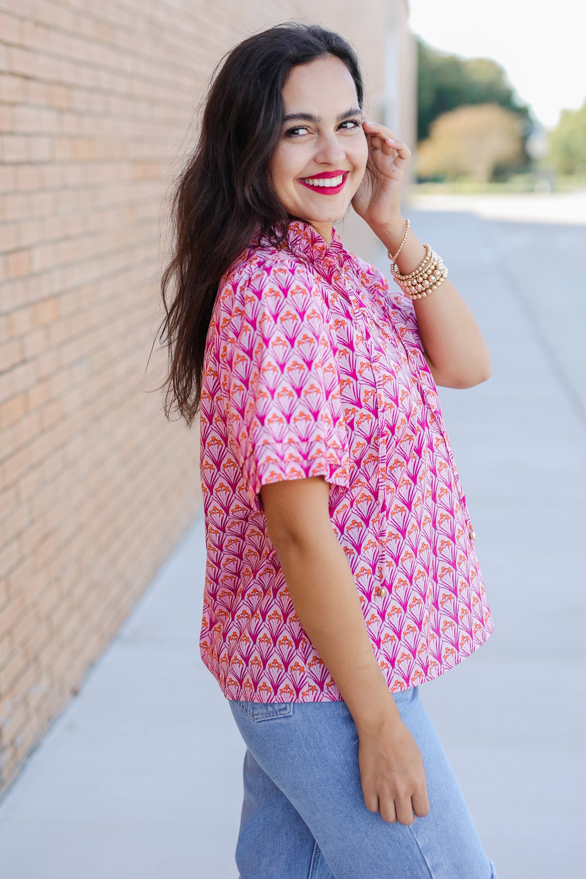 Woman wearing a pink patterned blouse and blue jeans standing against a brick wall.