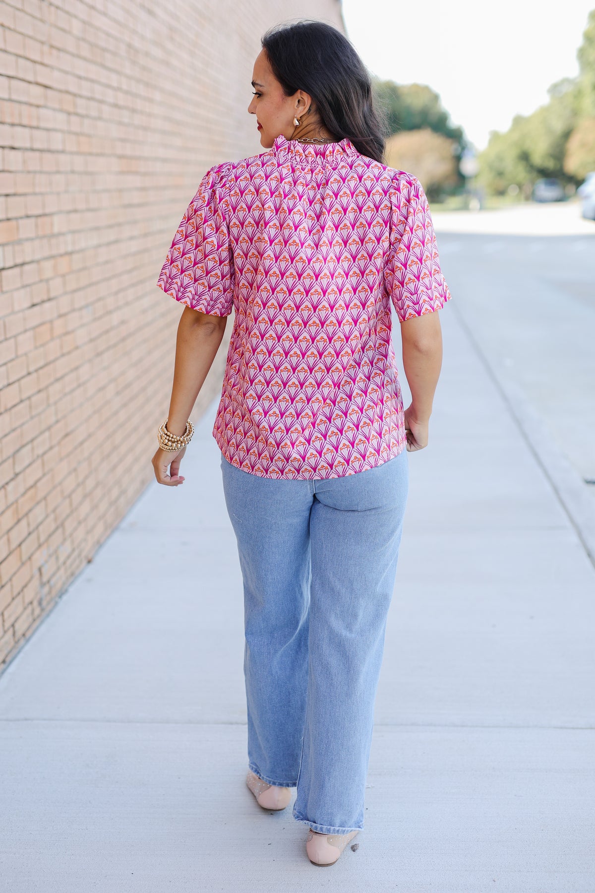 Woman wearing a pink patterned blouse and blue jeans walking on a sidewalk.