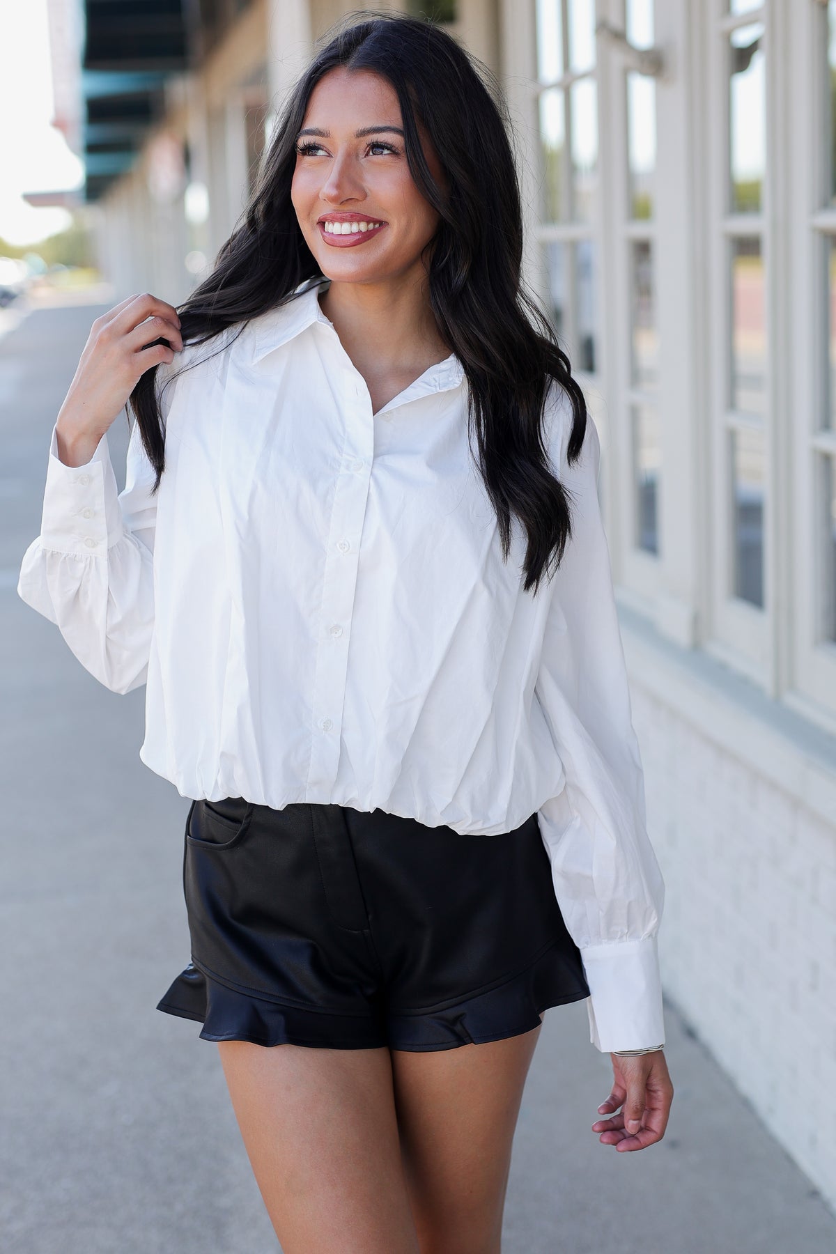 A woman wearing a white long-sleeve button-up blouse and black ruffled shorts, standing outdoors on a sidewalk next to a building with large windows.