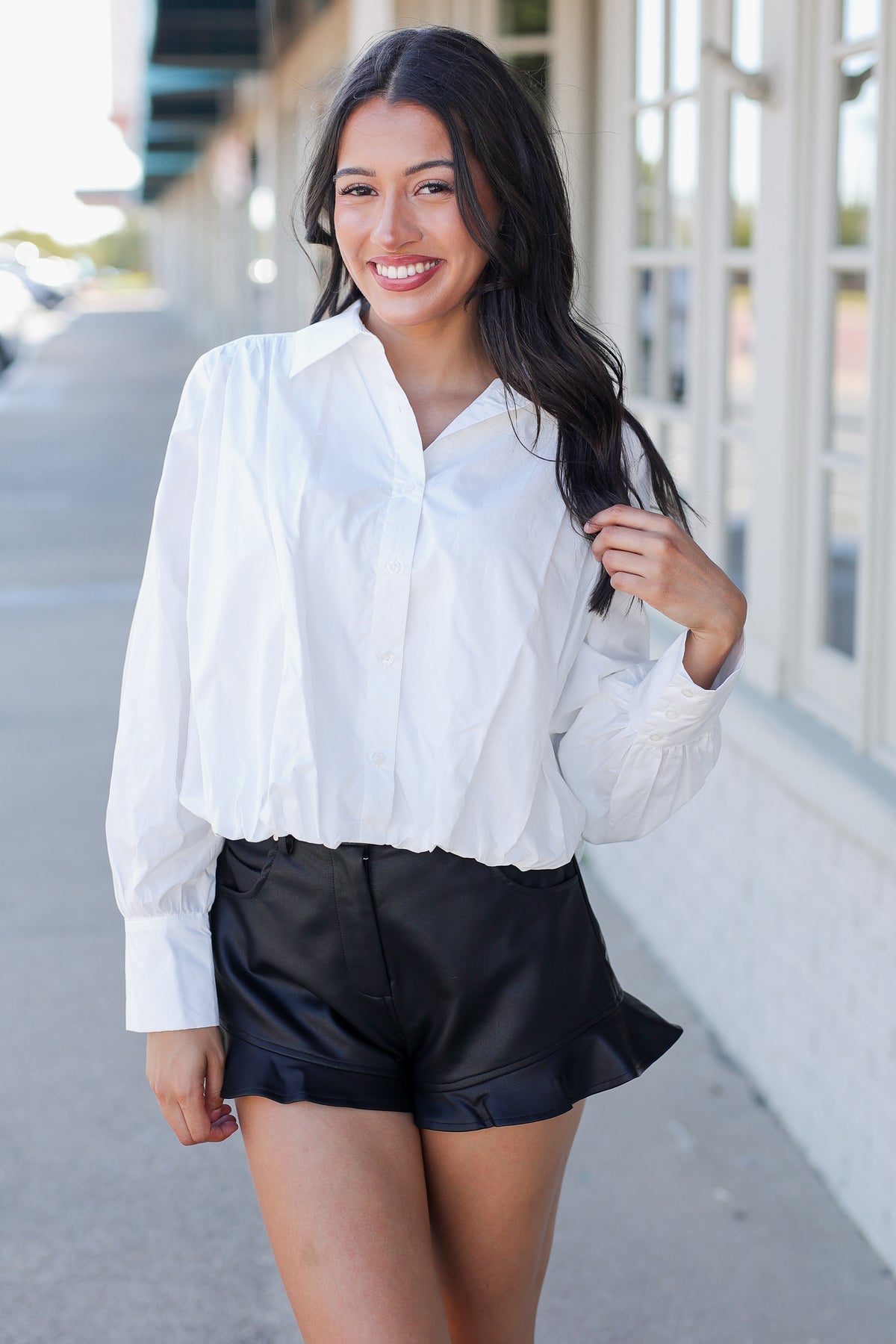 A woman wearing a white long-sleeve button-down shirt and black ruffled shorts, standing outdoors on a sidewalk next to a building with large windows.