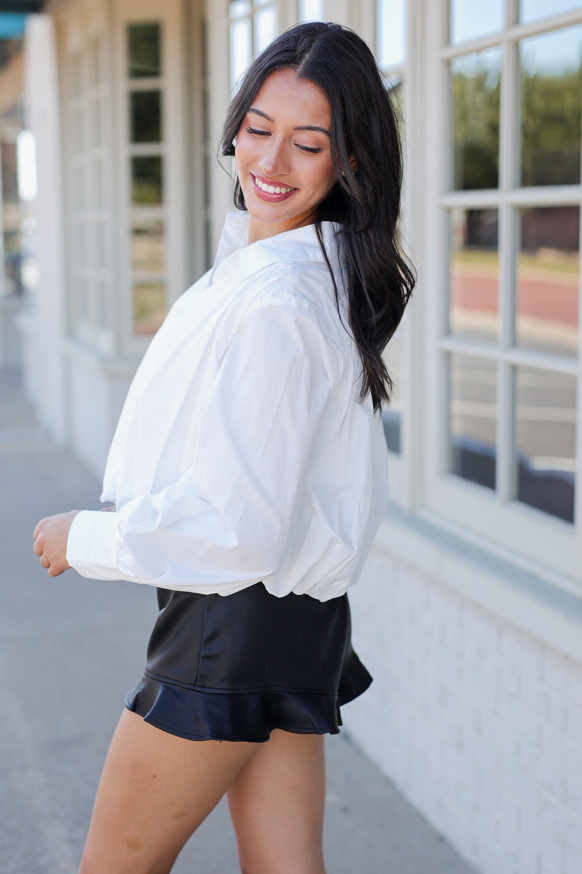 A woman wearing a white long-sleeve button-up shirt and black shorts, standing outdoors in front of a building with large windows.