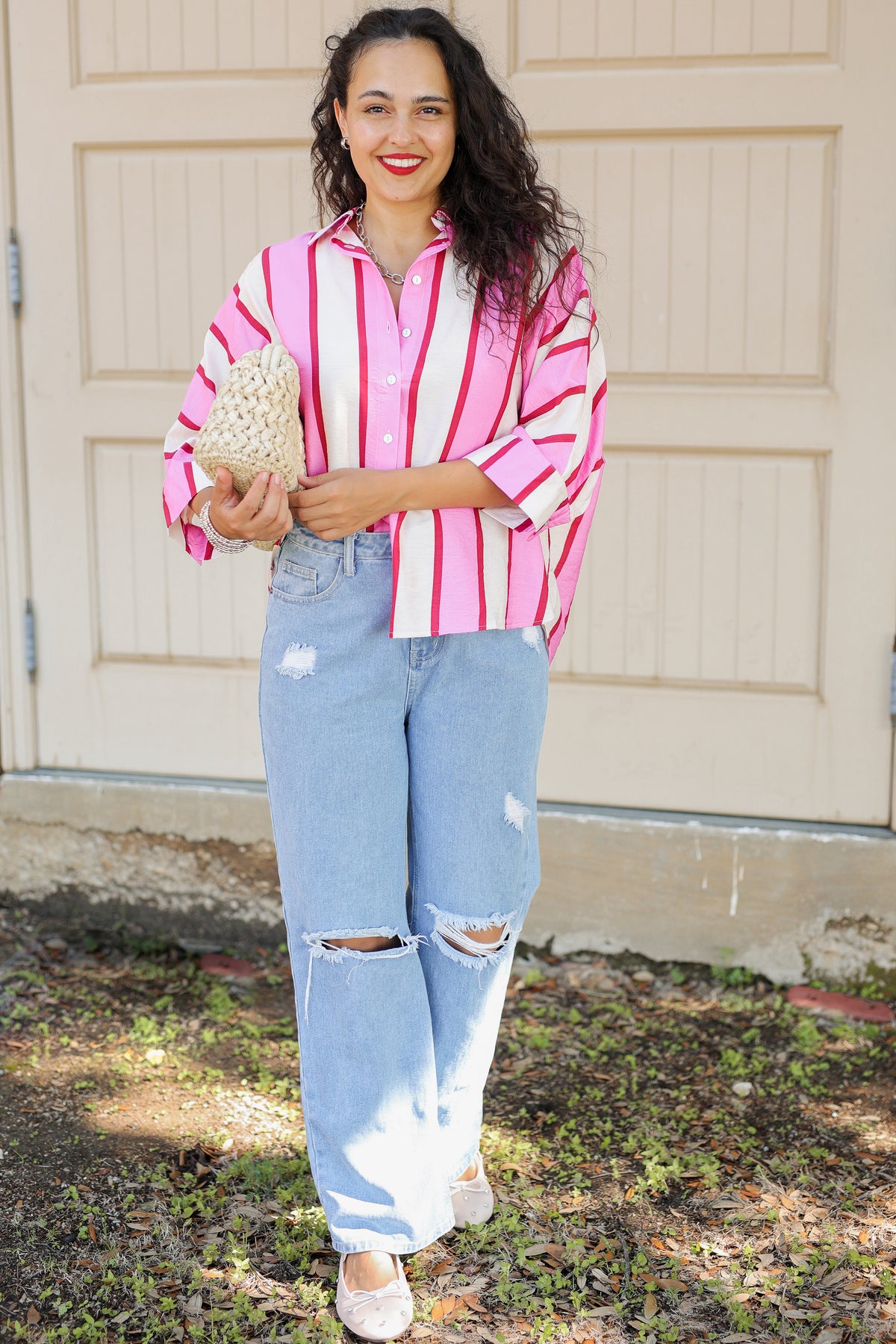 A woman stands outdoors in front of a beige garage door wearing a pink and white vertically striped button-down shirt, light-wash distressed jeans with ripped knees, and white flats. She is holding a woven straw clutch.