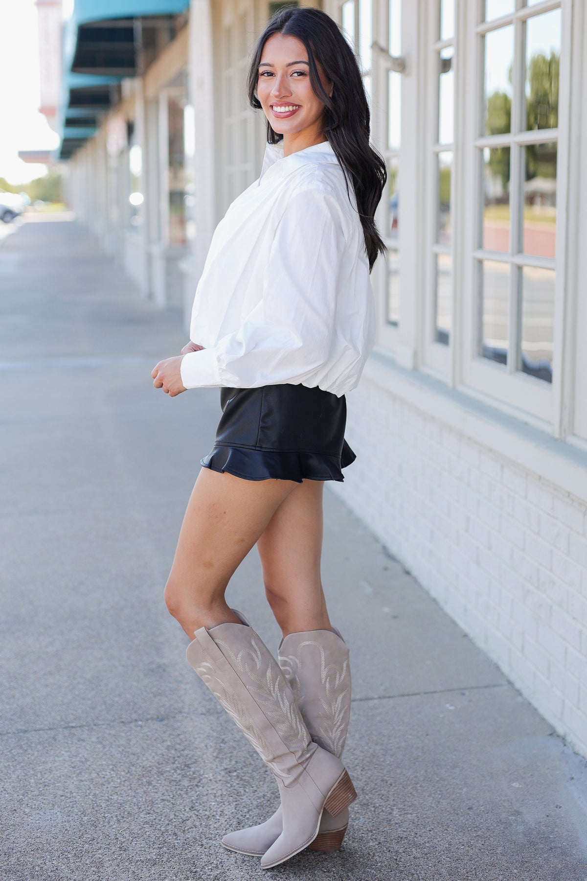 A woman posing on a city sidewalk wearing a white long-sleeve blouse, black ruffled shorts, and beige western-style knee-high boots.