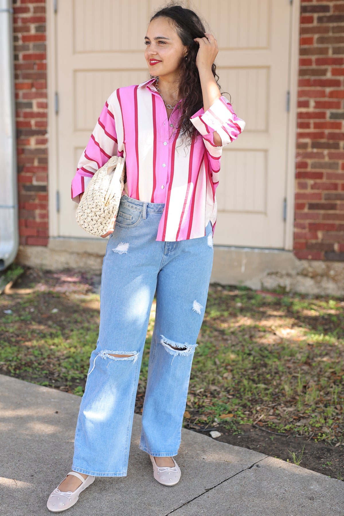 A woman stands outdoors in front of a brick building and beige garage door, wearing a pink and white vertically striped button-up shirt, light blue wide-leg distressed jeans, and white flats. She is holding a woven beige handbag.