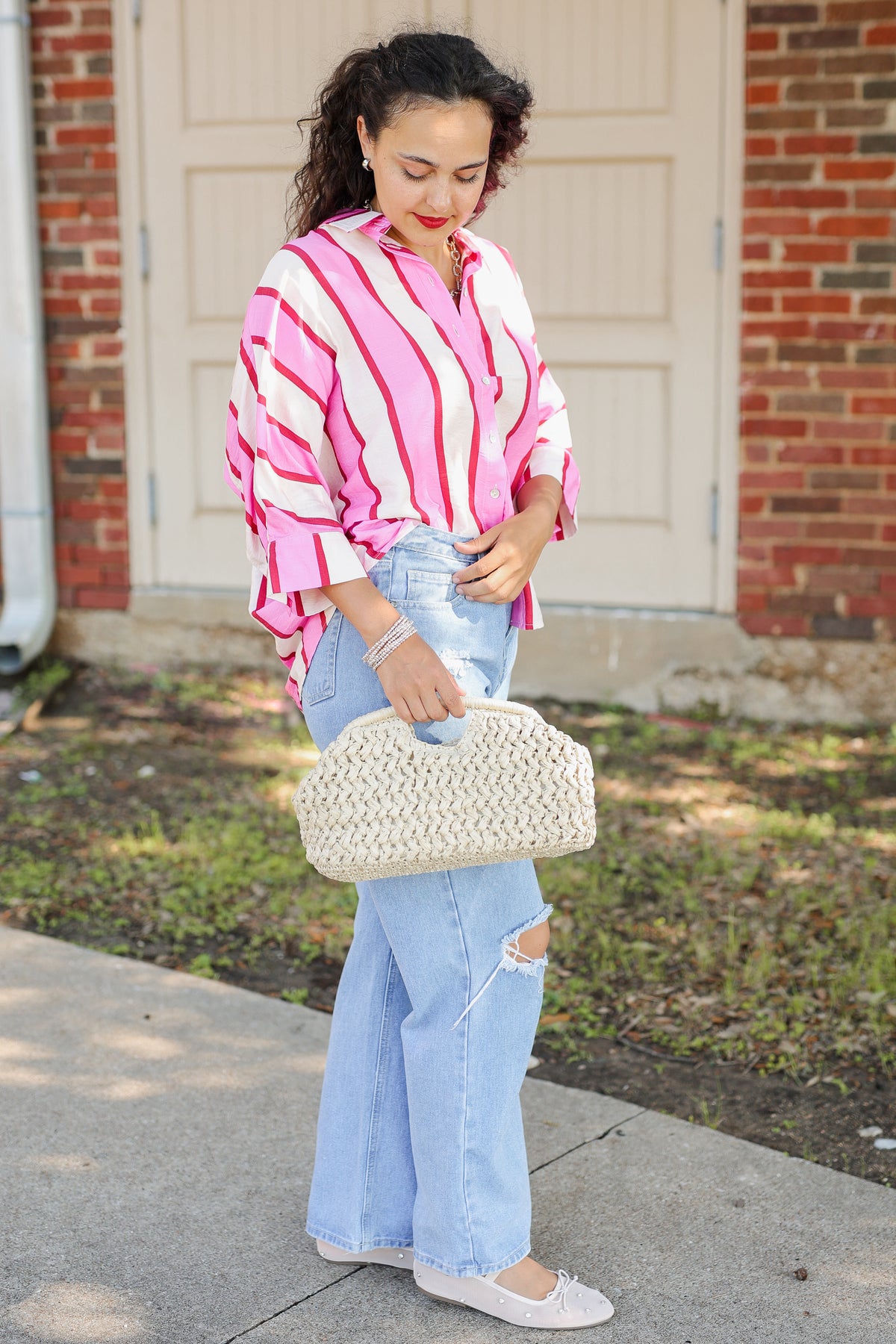 A woman stands outdoors in front of a brick wall and garage door, wearing a pink and white vertically striped button-up shirt, light blue wide-leg jeans, white flats, and holding a cream woven handbag.