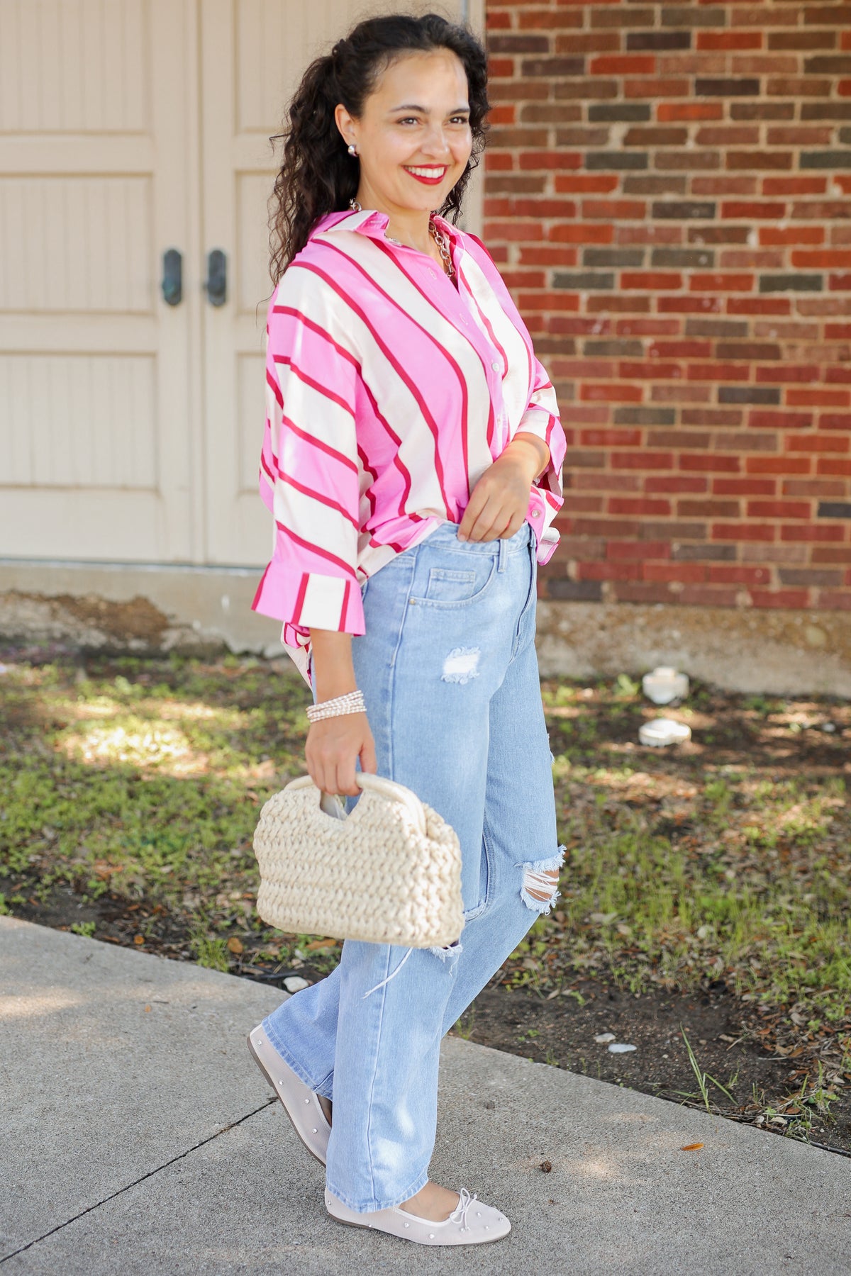 A woman stands outdoors on a sidewalk in front of a brick wall and garage door, wearing a pink and white striped long-sleeve blouse, light-wash distressed jeans, white flats, and holding a cream woven handbag.