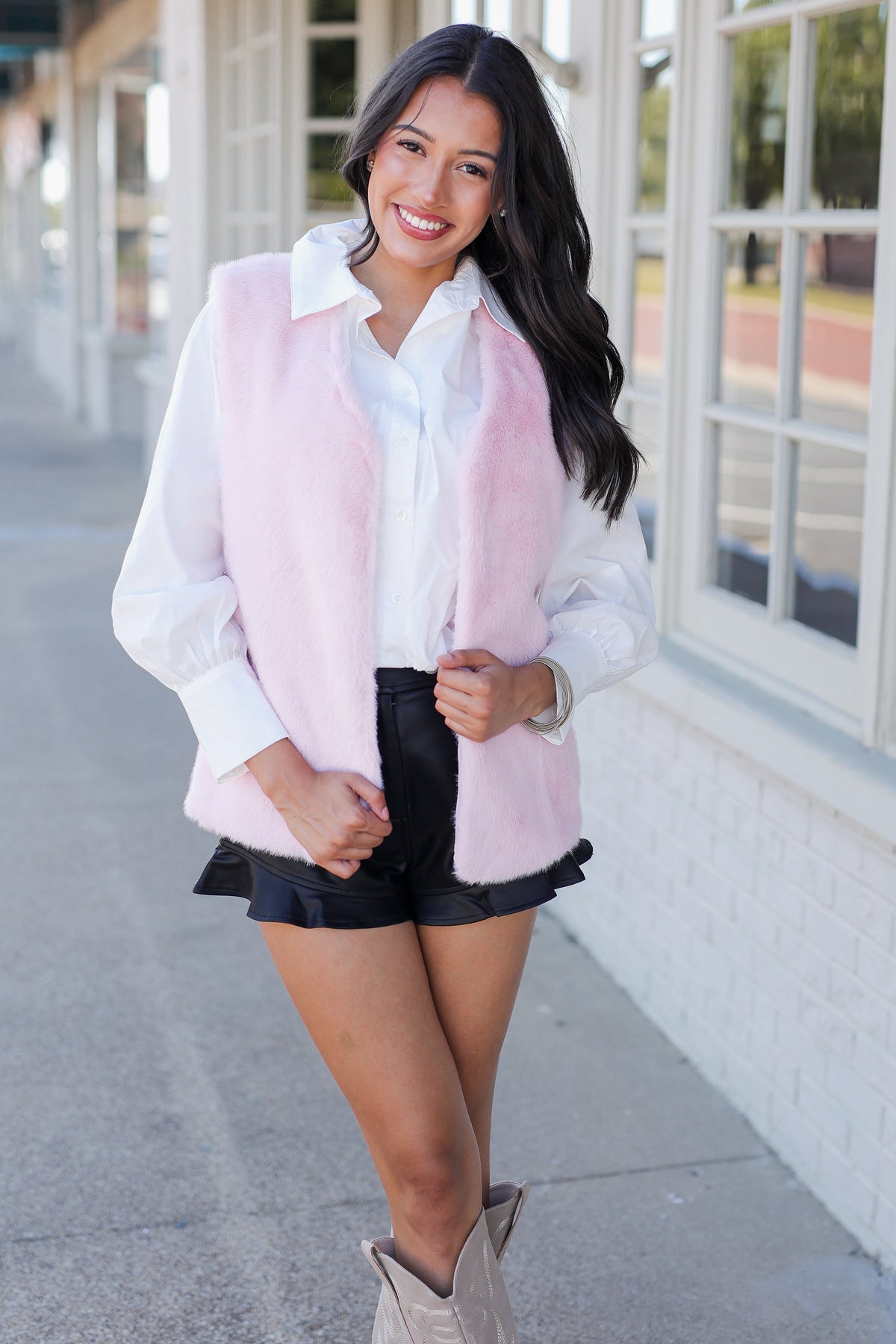 A woman modeling a light pink faux fur vest over a white collared shirt, paired with black shorts and beige cowboy boots, standing on a sidewalk next to a white building.