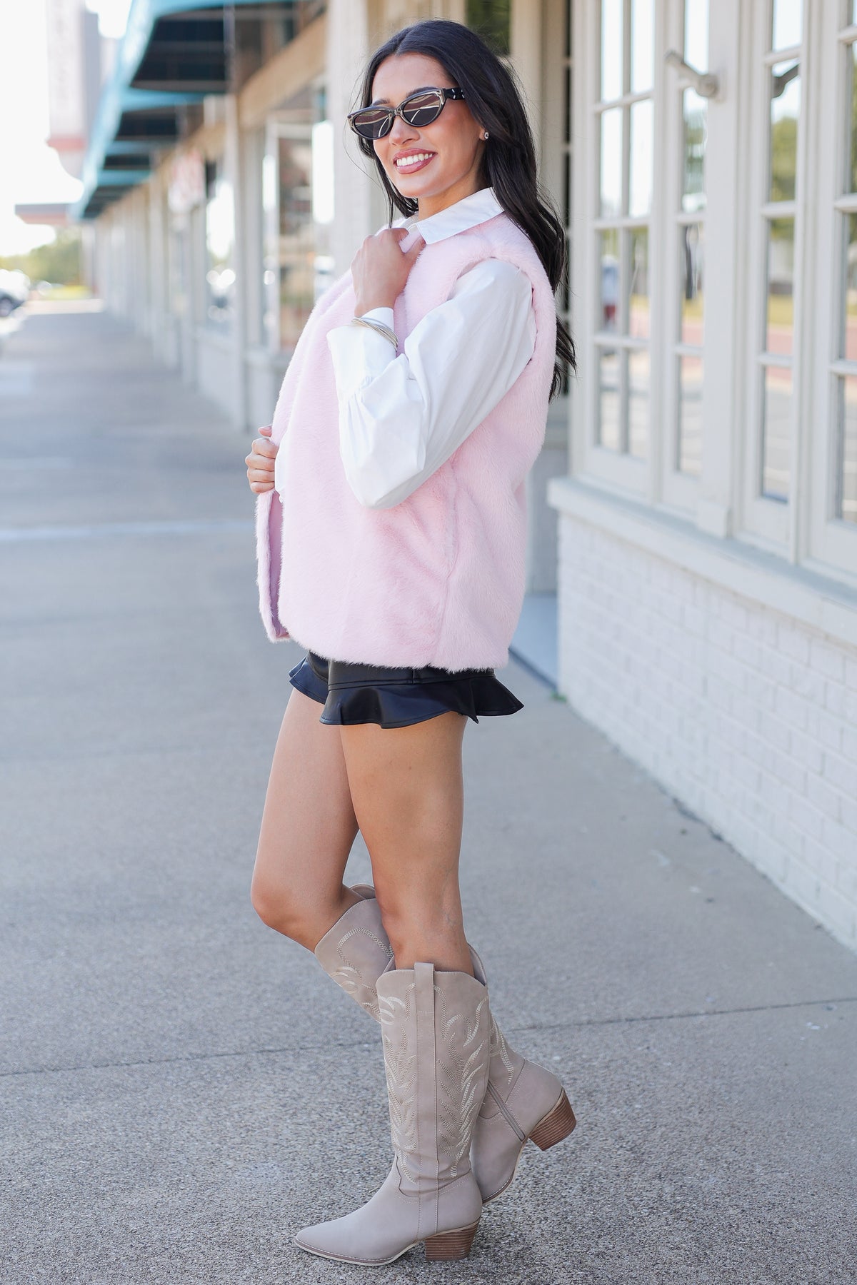 A woman wearing a light pink faux-fur vest over a white collared shirt, black ruffled shorts, and beige knee-high boots, standing on a sidewalk next to a building.