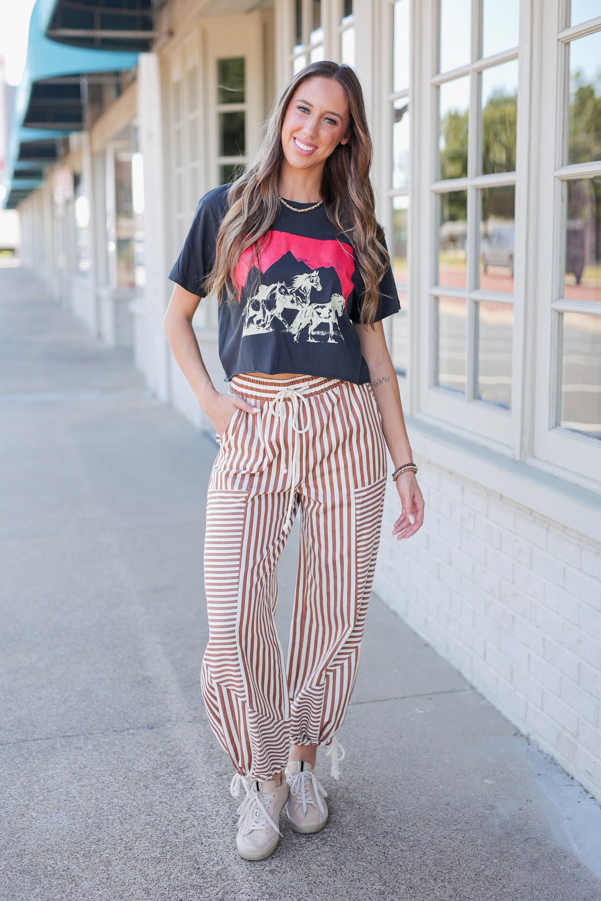 A woman stands on a sidewalk wearing a dark graphic t-shirt with a pink and white horse design, paired with brown and white vertically striped drawstring pants and white sneakers.