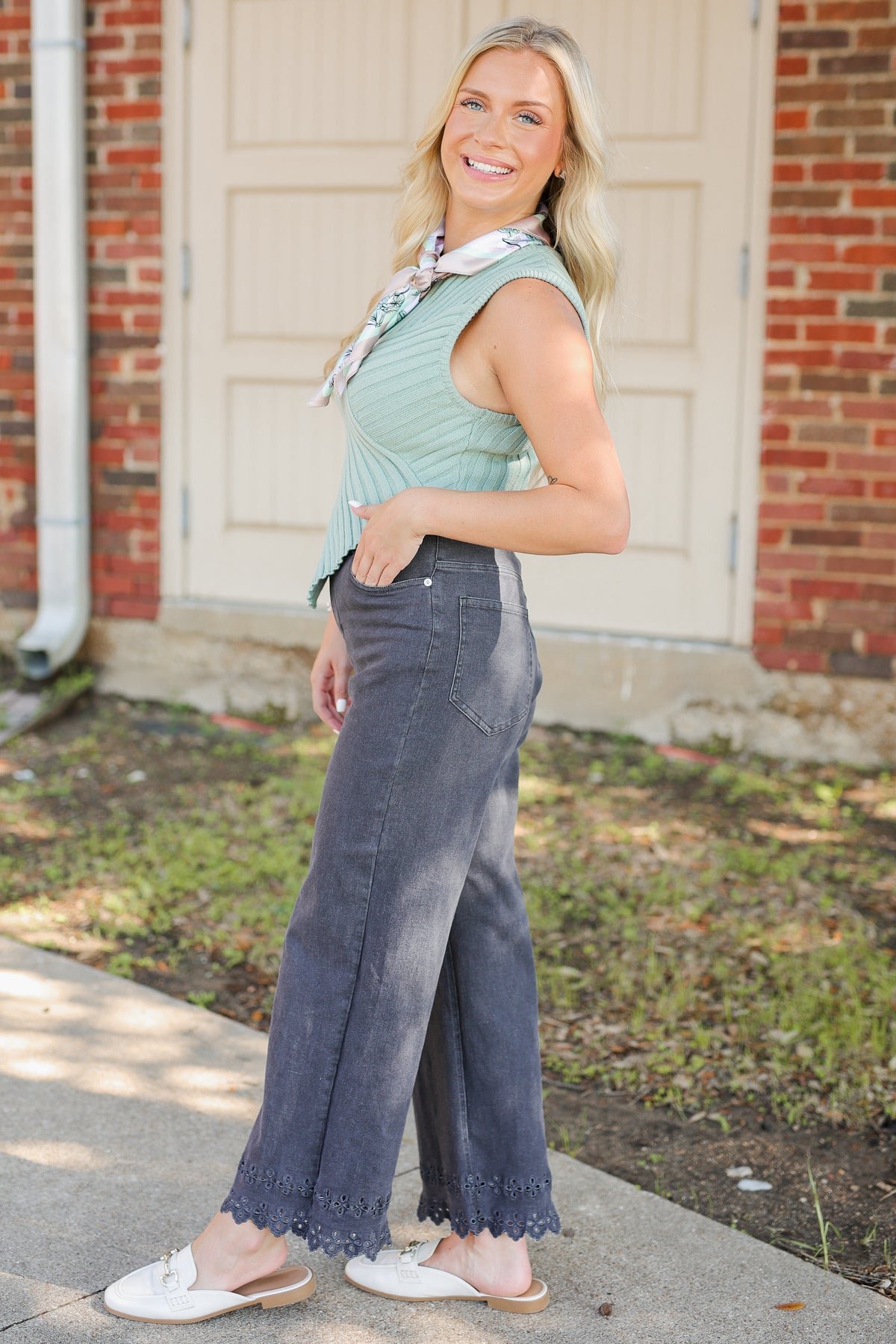 A woman stands outdoors in front of a brick building and white door, wearing a light sage green ribbed sleeveless top, dark gray wide-leg jeans with scalloped hems, and white slide sandals.