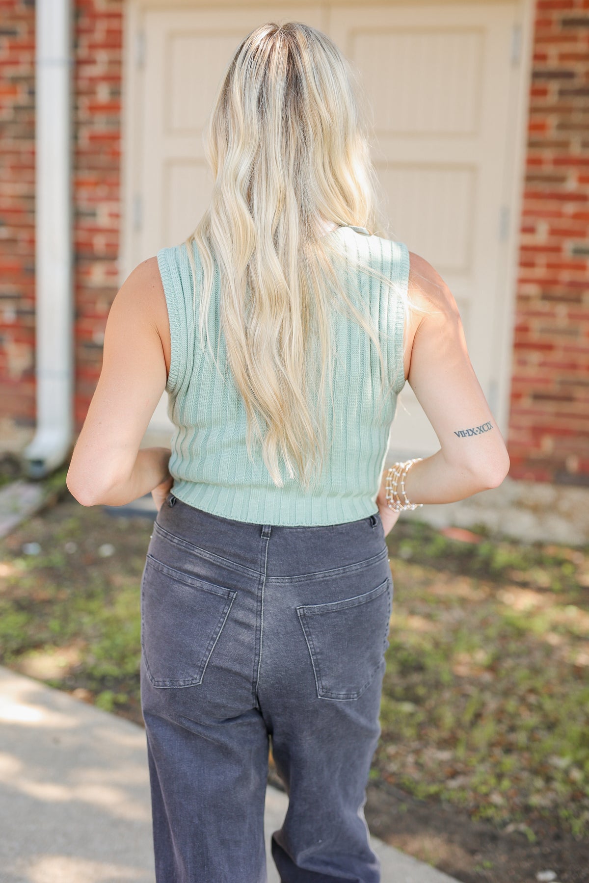 A woman viewed from behind wearing a sage green ribbed sleeveless top and dark gray jeans, standing outdoors in front of a brick house door.