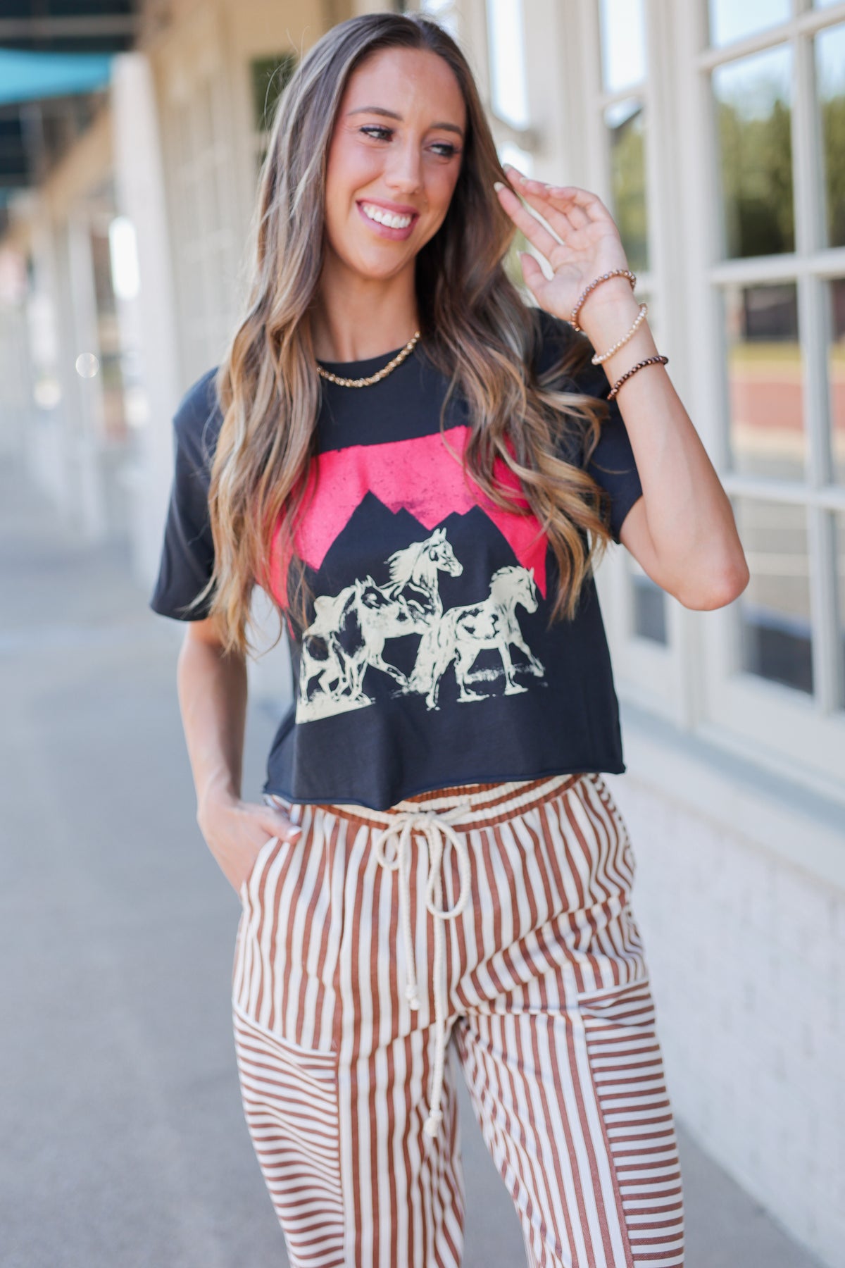 A woman stands outdoors on a sidewalk next to a building, wearing loose brown and white vertically striped drawstring pants, a black graphic t-shirt, and beige sneakers.