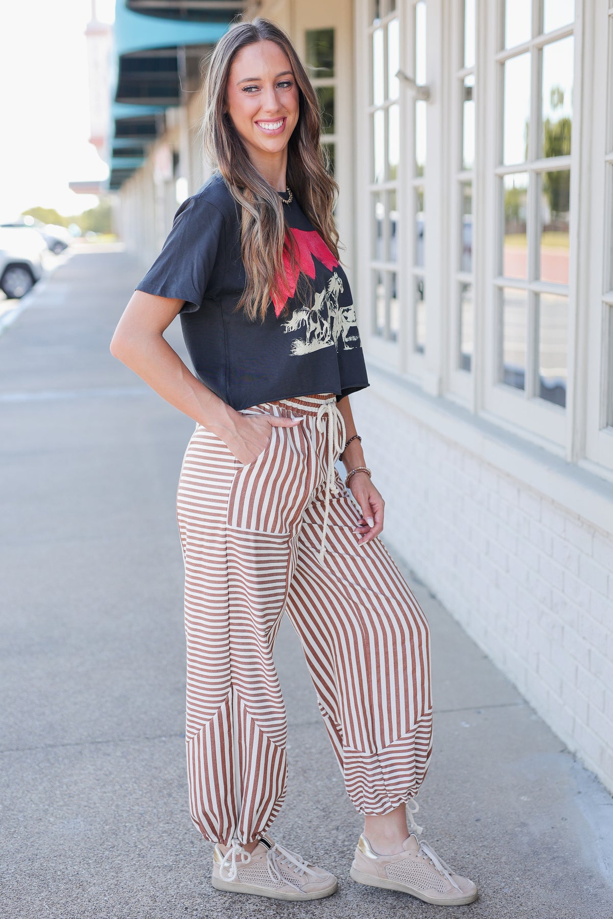 A woman stands outdoors on a sidewalk next to a building, wearing loose brown and white vertically striped drawstring pants, a black graphic t-shirt, and beige sneakers.