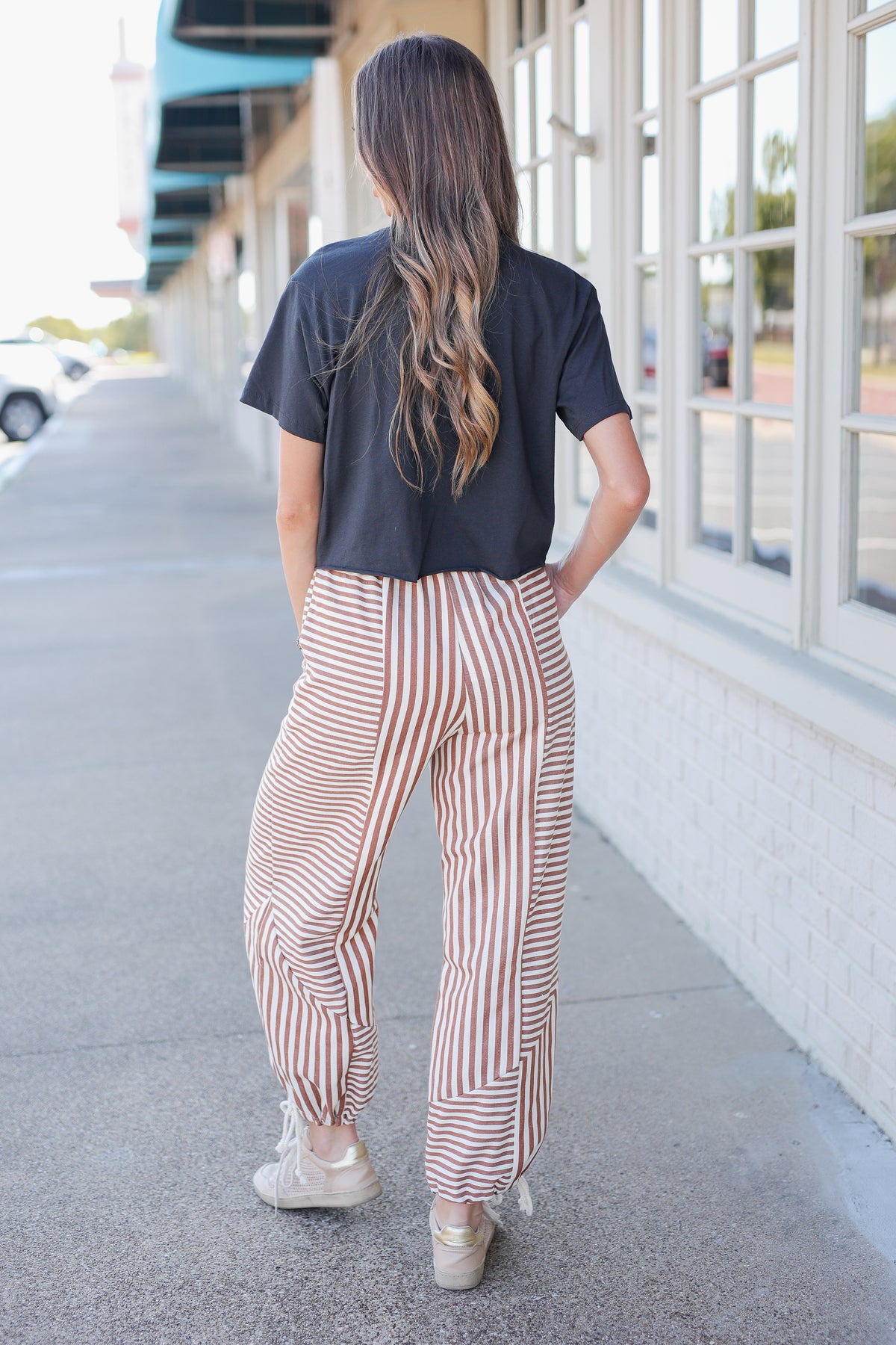 A woman viewed from behind wearing a black t-shirt and high-waisted, wide-leg striped pants in beige and brown, standing on a sidewalk next to a building.