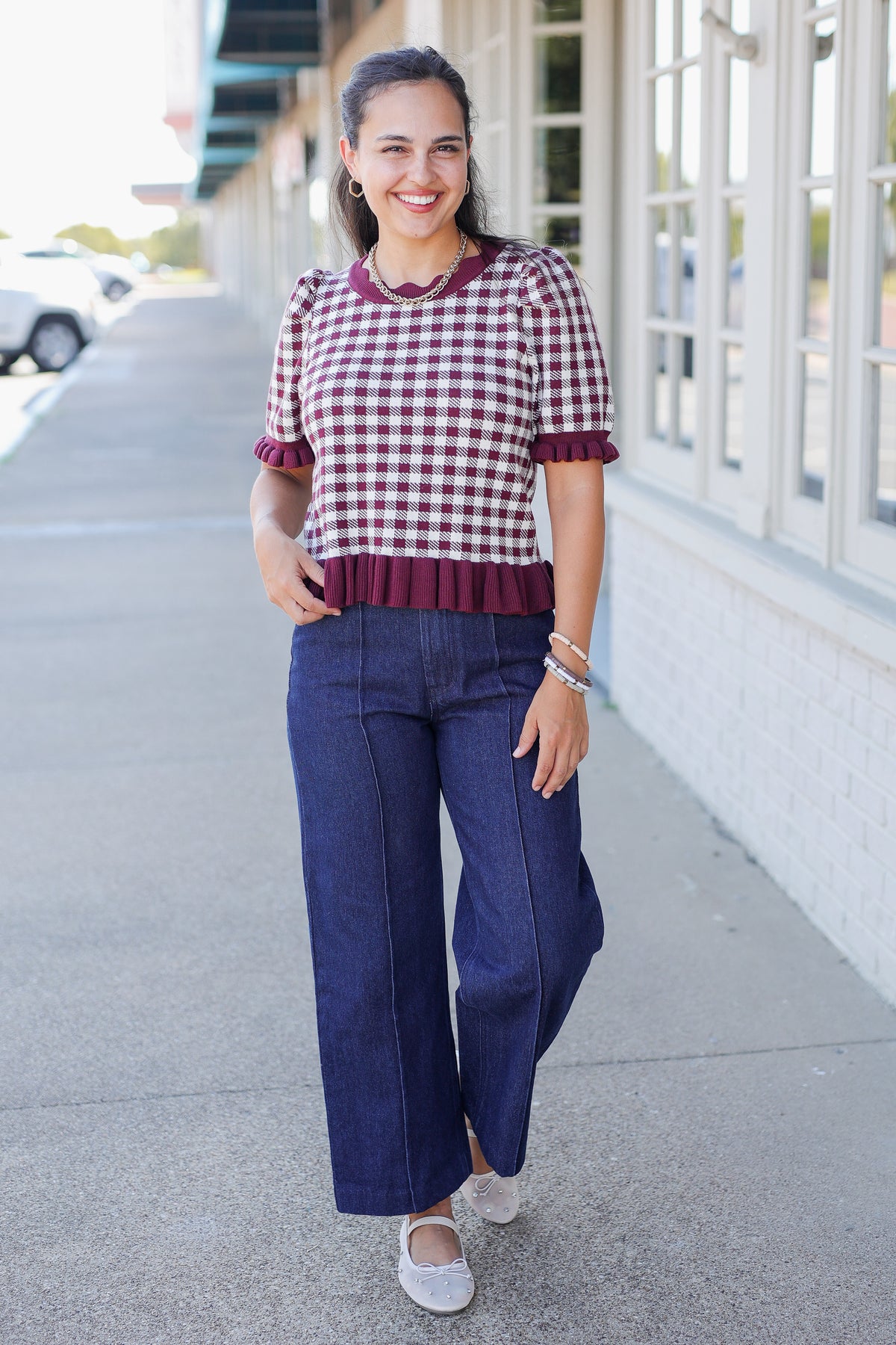 A woman stands on a sidewalk wearing a short-sleeve gingham top with dark red ruffled trim and dark blue wide-leg jeans.