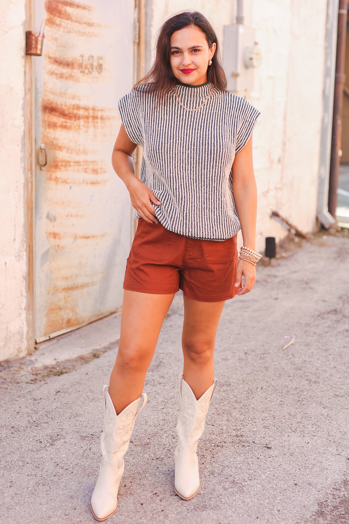 Woman wearing a striped top, rust-colored shorts, and white cowboy boots standing in an urban setting.
