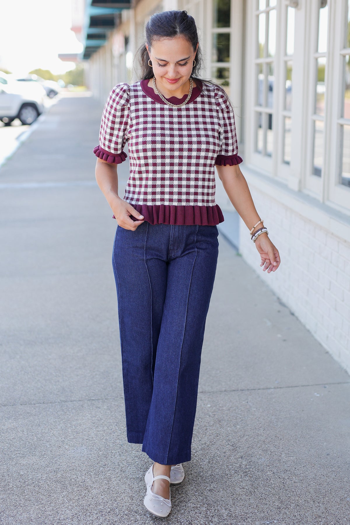 A woman walking outdoors on a sidewalk wearing a short-sleeve burgundy and white gingham top with ruffled trim, paired with dark blue wide-leg pants and white espadrille flats.