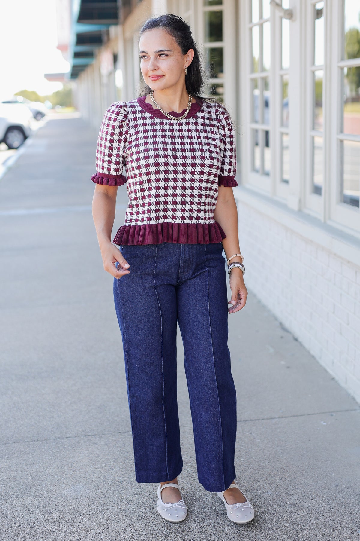 A woman stands on a sidewalk wearing a short-sleeve red and white gingham top with a dark red ruffled hem, paired with dark blue wide-leg jeans and white flats.