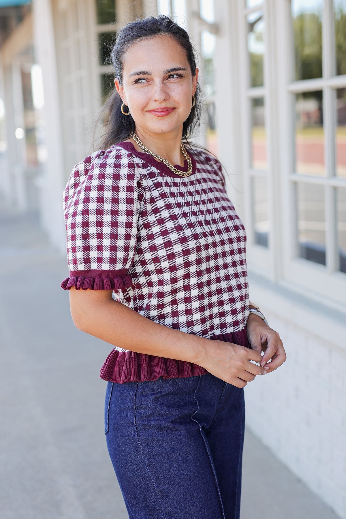 A woman modeling a short-sleeve top with a burgundy and white gingham pattern and ruffled trim, paired with dark blue jeans, standing in front of a white building with large windows.