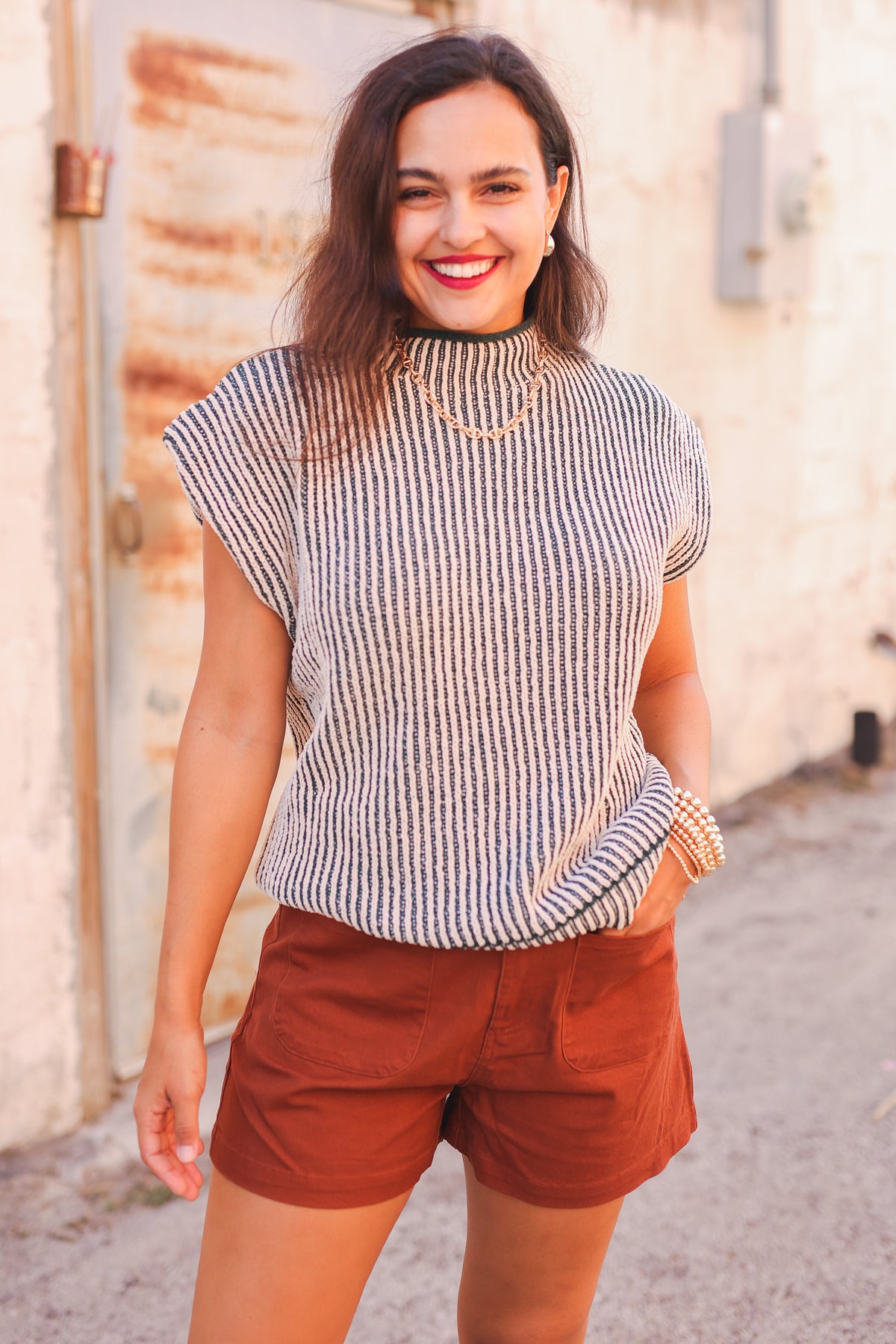 Woman wearing a striped sleeveless top and brown shorts standing against a rustic wall.