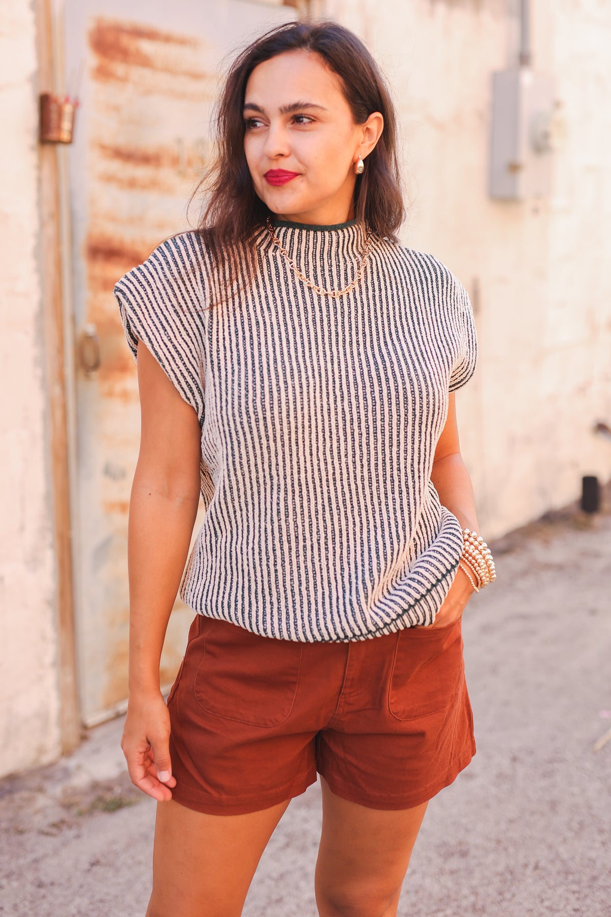 Woman wearing a striped sleeveless top and brown shorts against a rustic wall.