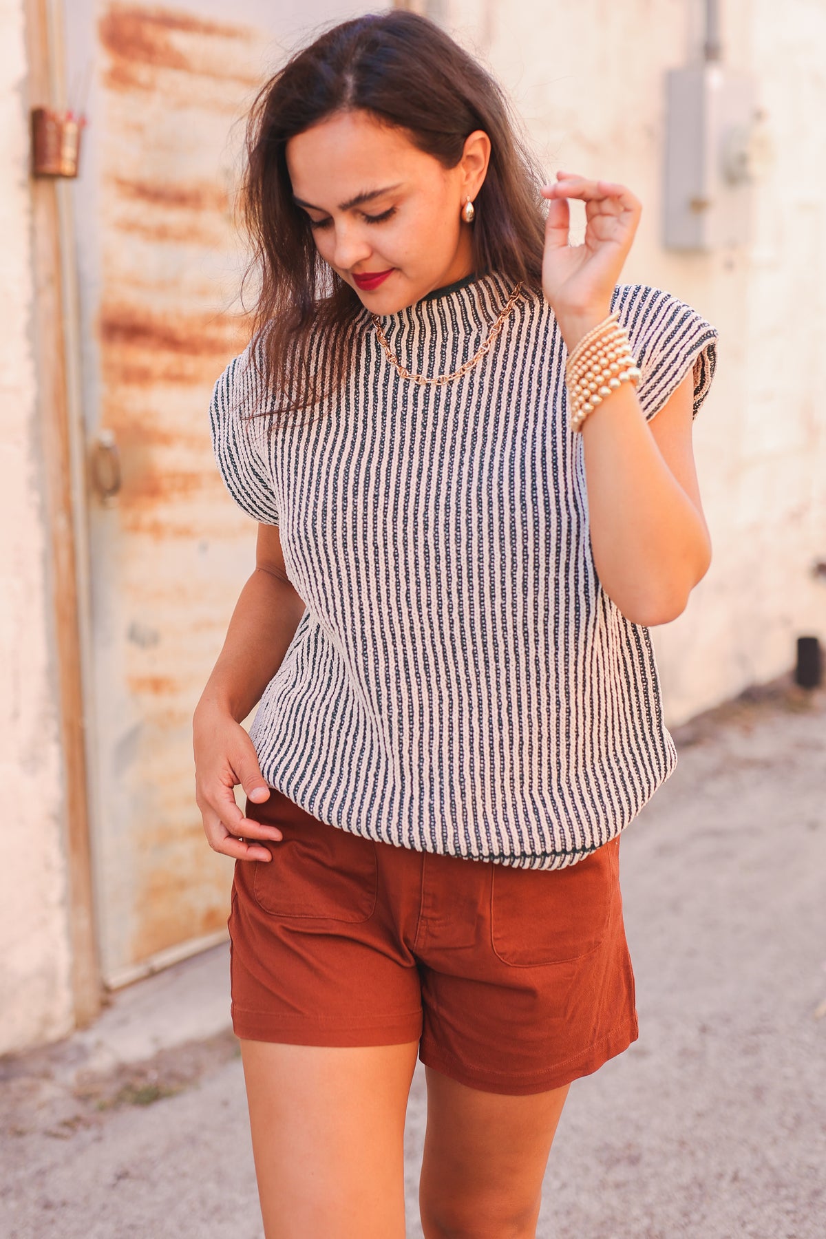 Woman wearing a striped shirt and brown shorts standing in front of a rustic wall.