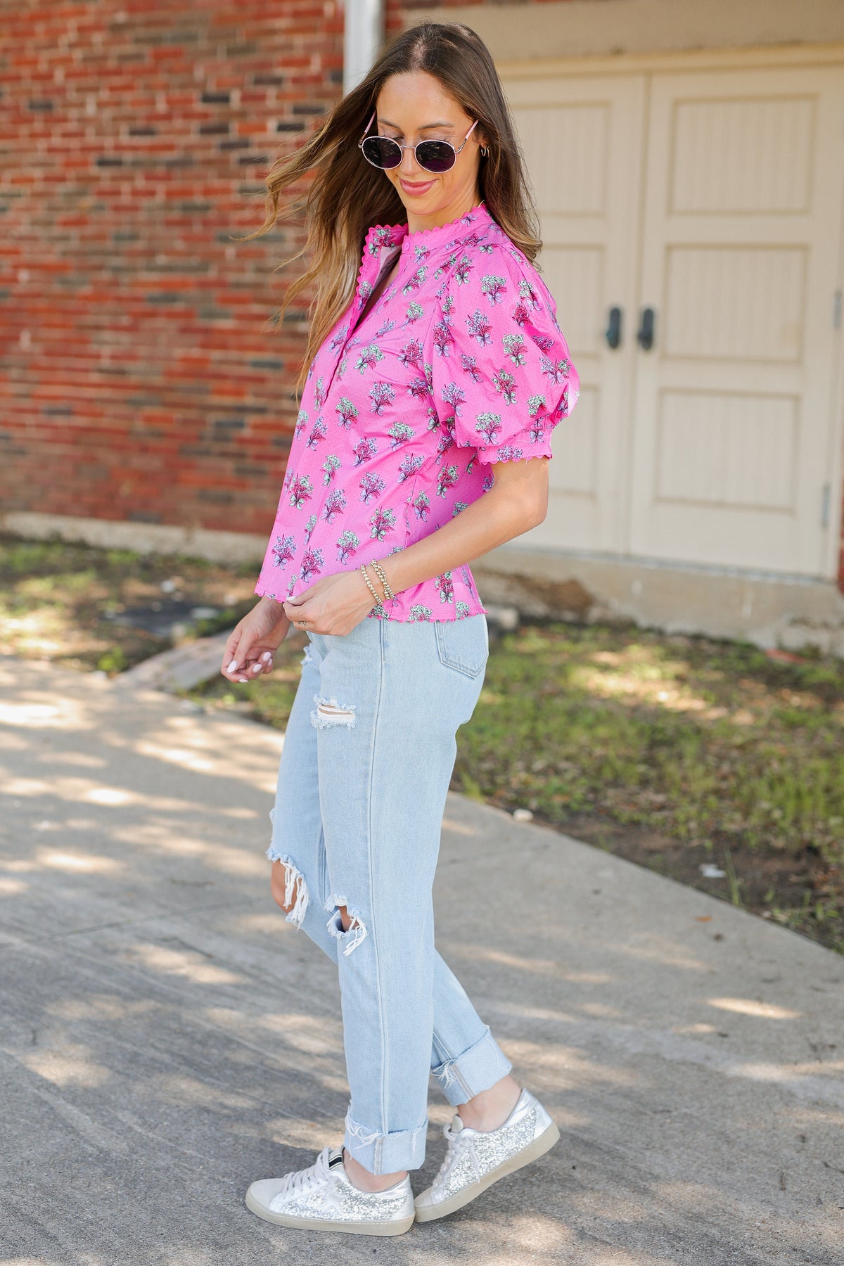 A woman wearing a pink short-sleeve button-up shirt with a green and pink floral pattern, light-wash distressed jeans, and silver sneakers, standing outdoors in front of a brick wall and garage door.