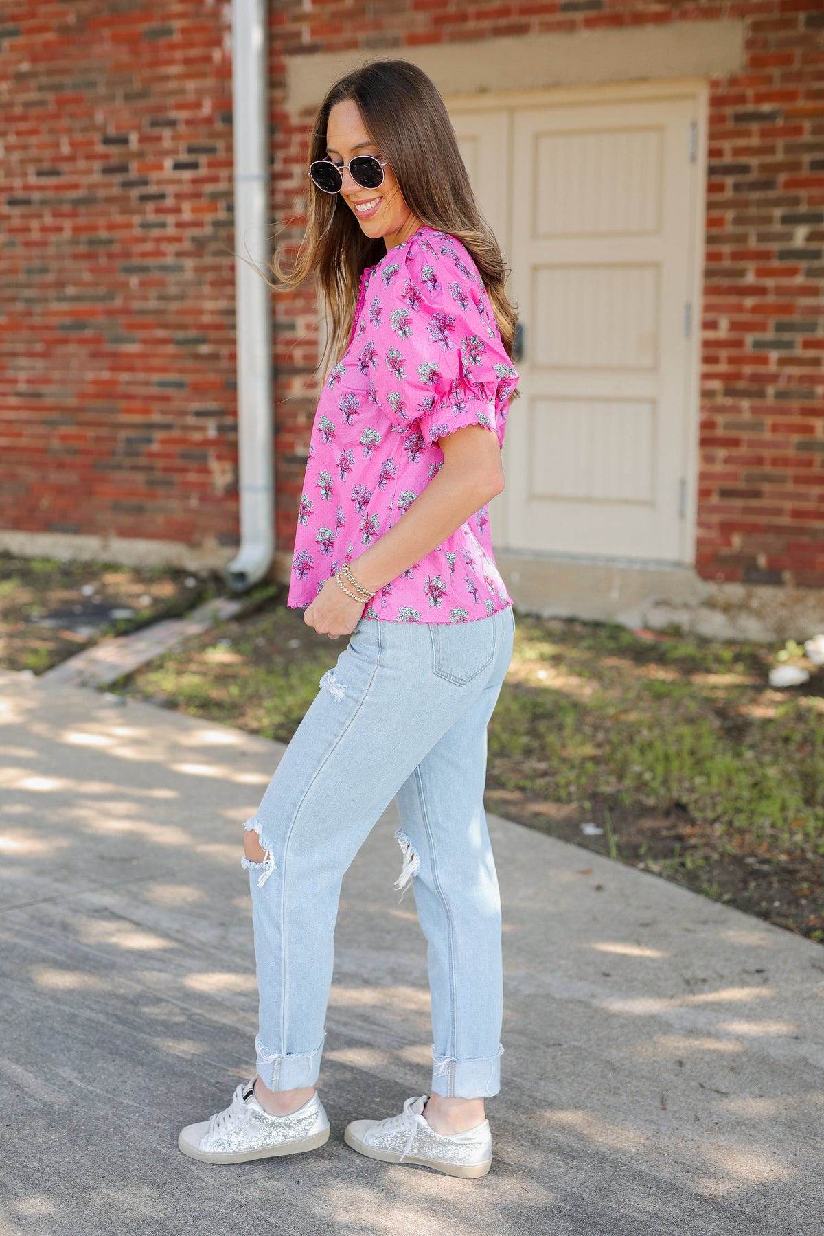 A woman wearing a pink short-sleeve blouse with a black floral pattern, light-wash distressed jeans, and white sneakers, standing outdoors in front of a brick building.