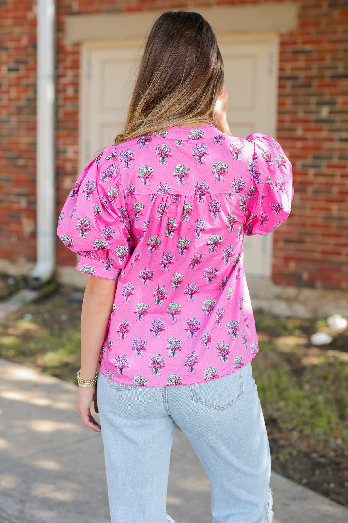 A woman viewed from behind wearing a bright pink short-sleeve blouse with a green and purple floral pattern, paired with light blue jeans, standing outdoors in front of a brick building.