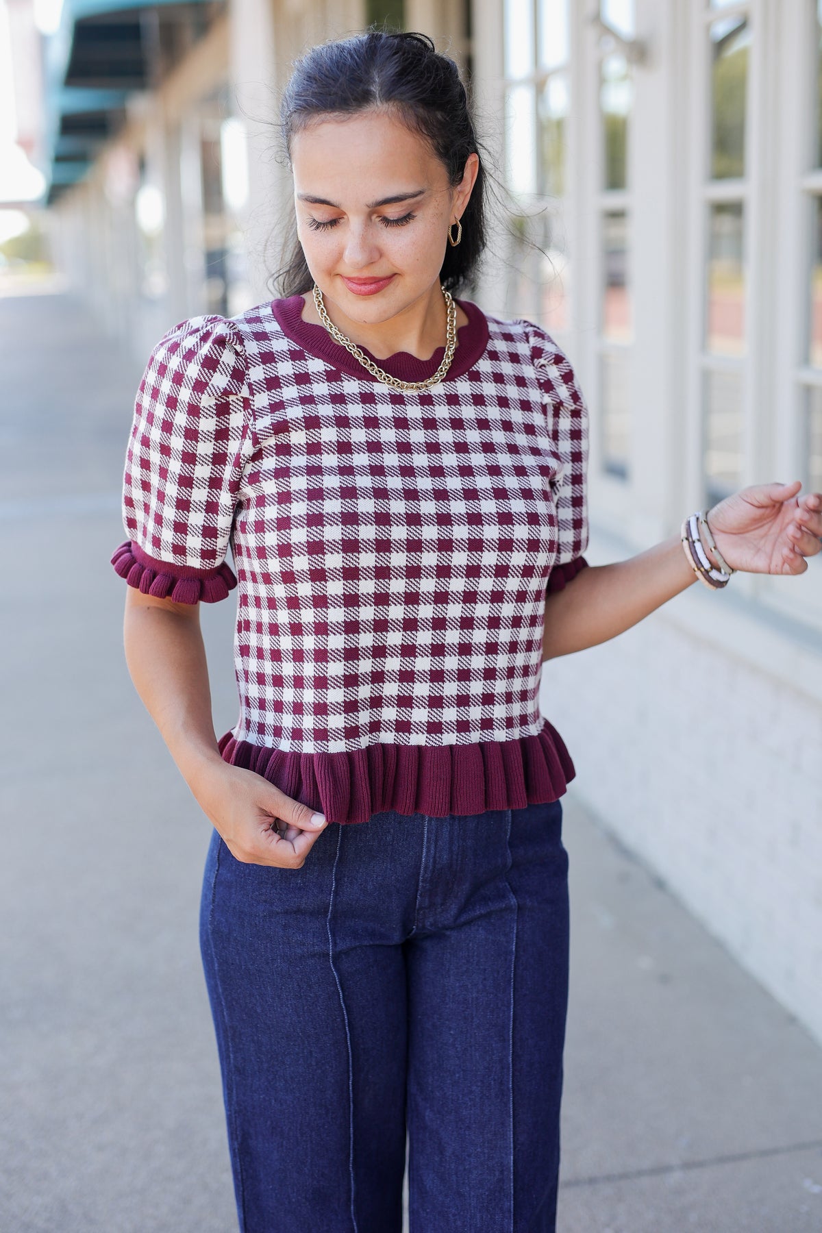 A woman wearing a burgundy and white gingham short-sleeve top with a ruffled hem and dark blue jeans, standing outdoors on a sidewalk.