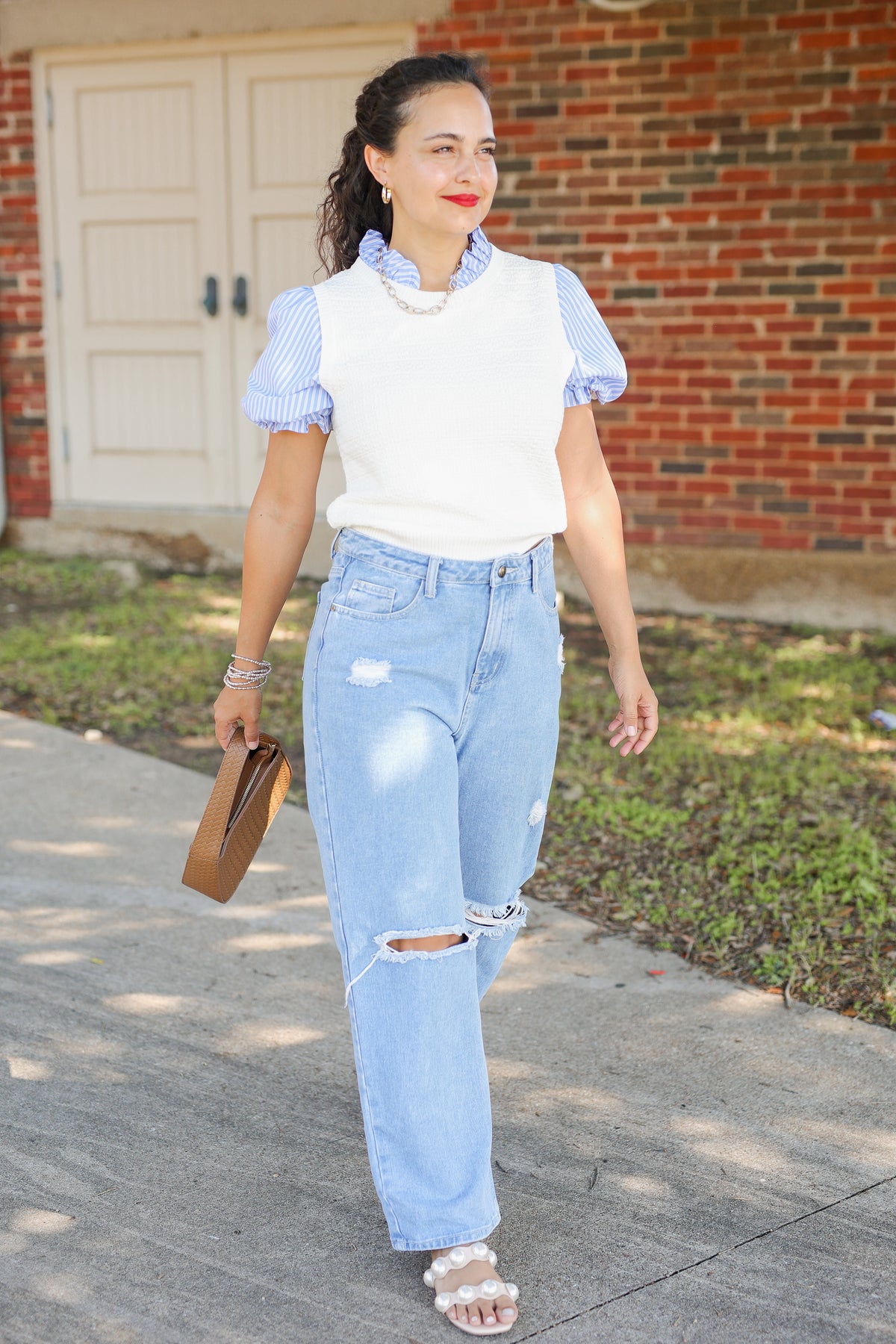 A woman walking outdoors on a sidewalk in front of a brick building, wearing a white sleeveless top over light blue and white gingham short-sleeve blouse, light-wash distressed jeans, and white slide sandals, while holding a brown clutch.
